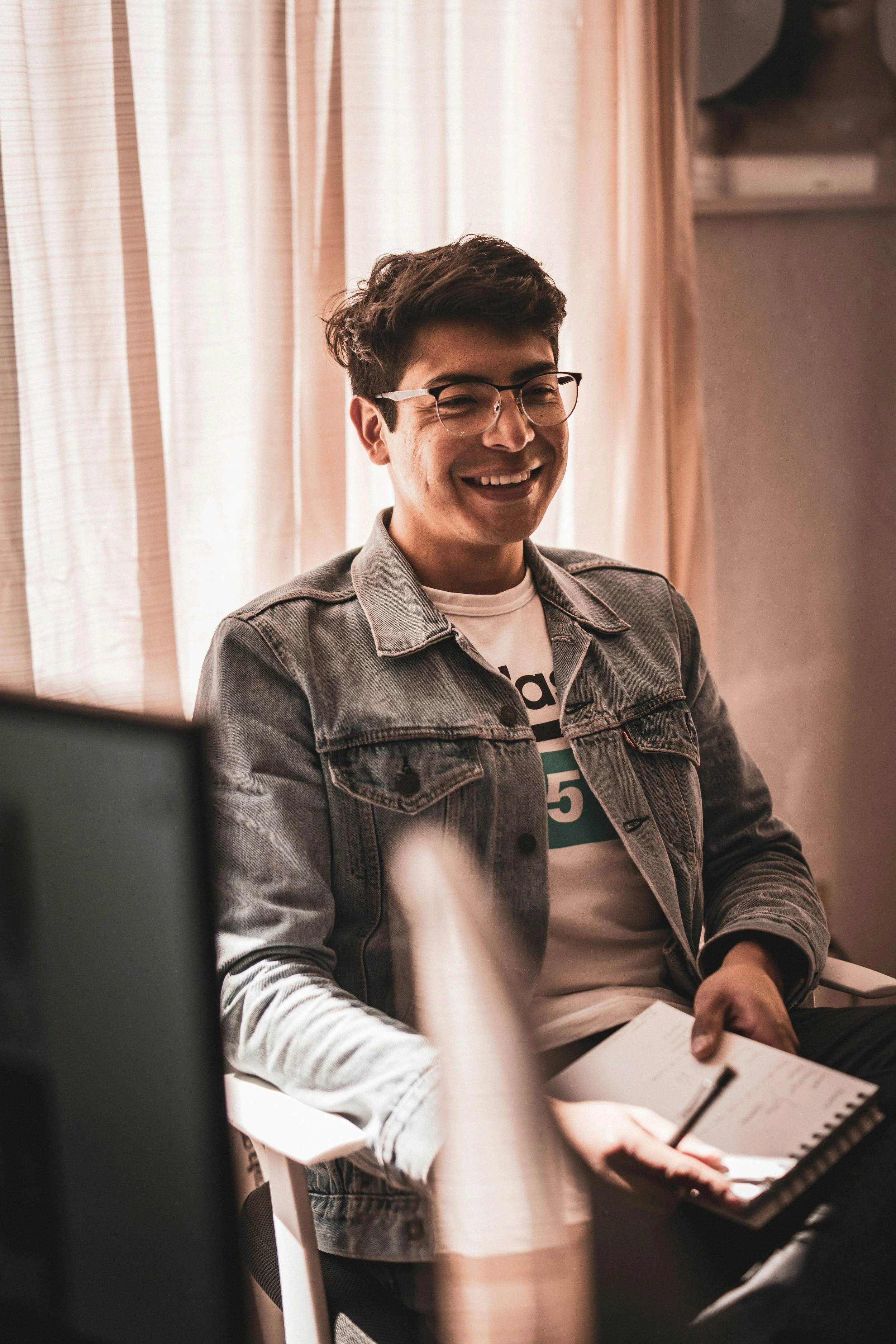 A young man with glasses and dark hair, wearing a denim jacket and a t-shirt, smiling and looking slightly downward while sitting at a desk with a notebook and pen in hand, in a well-lit room with curtains in the background.