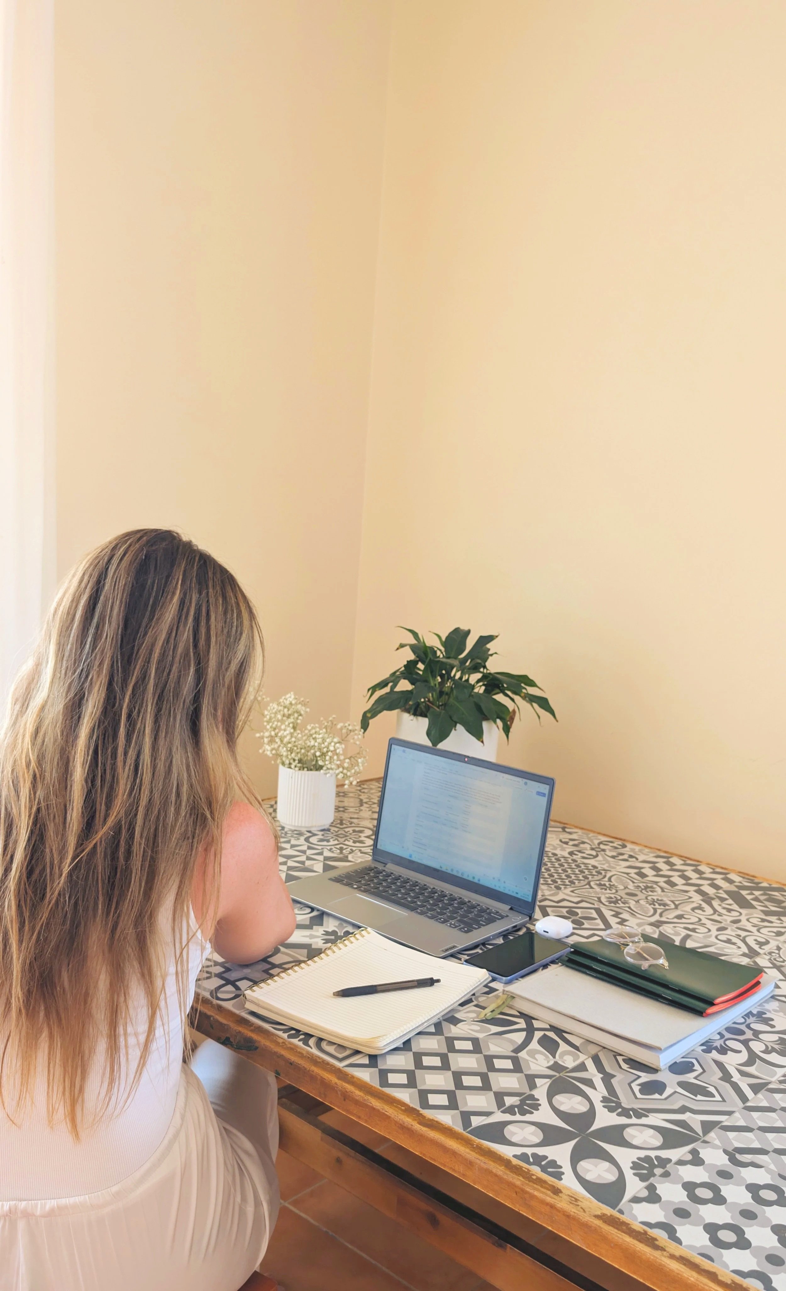 A woman with long blonde hair works on a laptop at a patterned table. There are notebooks, a smartphone, and glasses on the table. In the background, there are two potted plants and a beige wall.