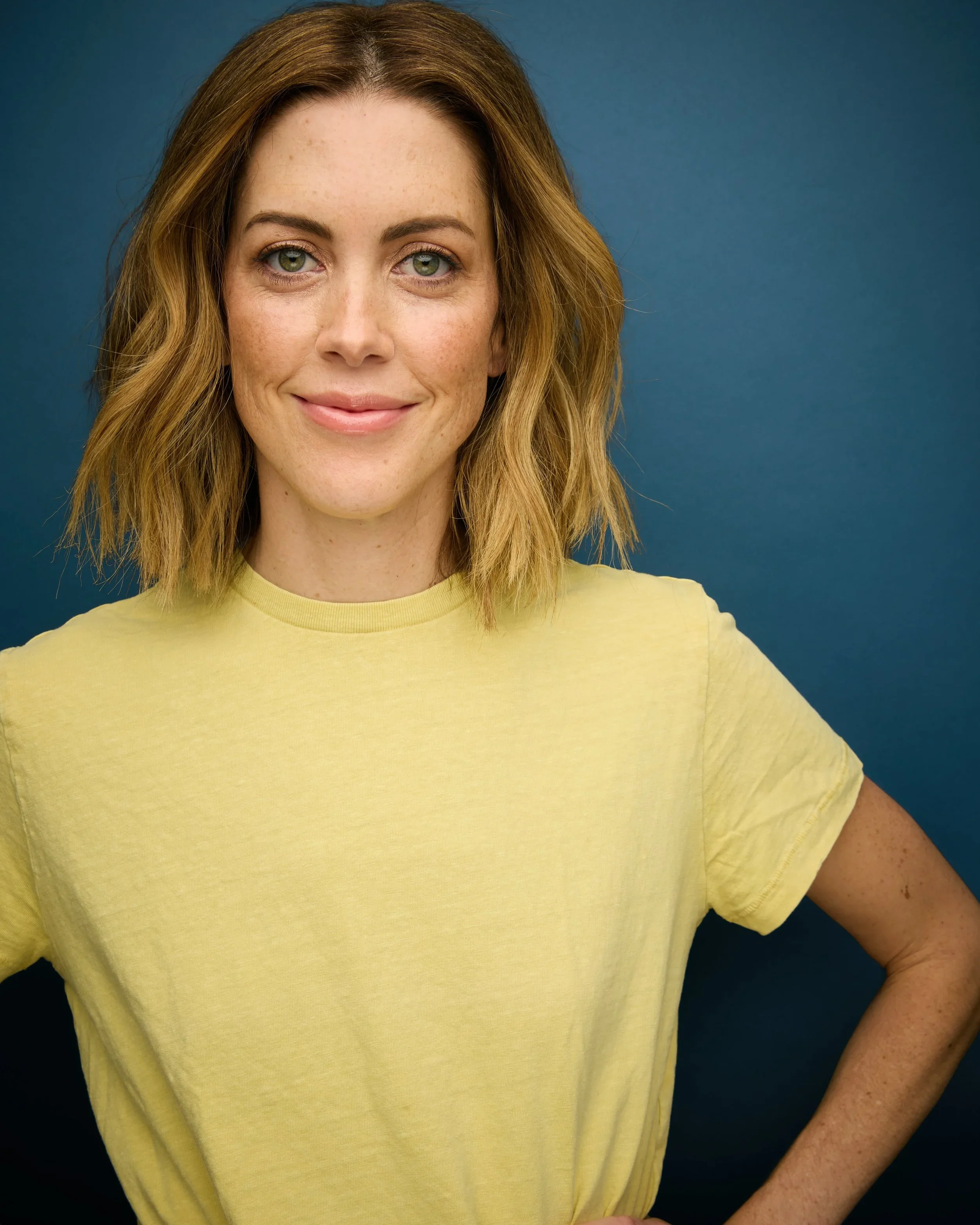 Close-up of a woman with wavy light brown hair, green eyes, light skin with freckles, smiling, wearing a yellow T-shirt, standing against a solid blue background.