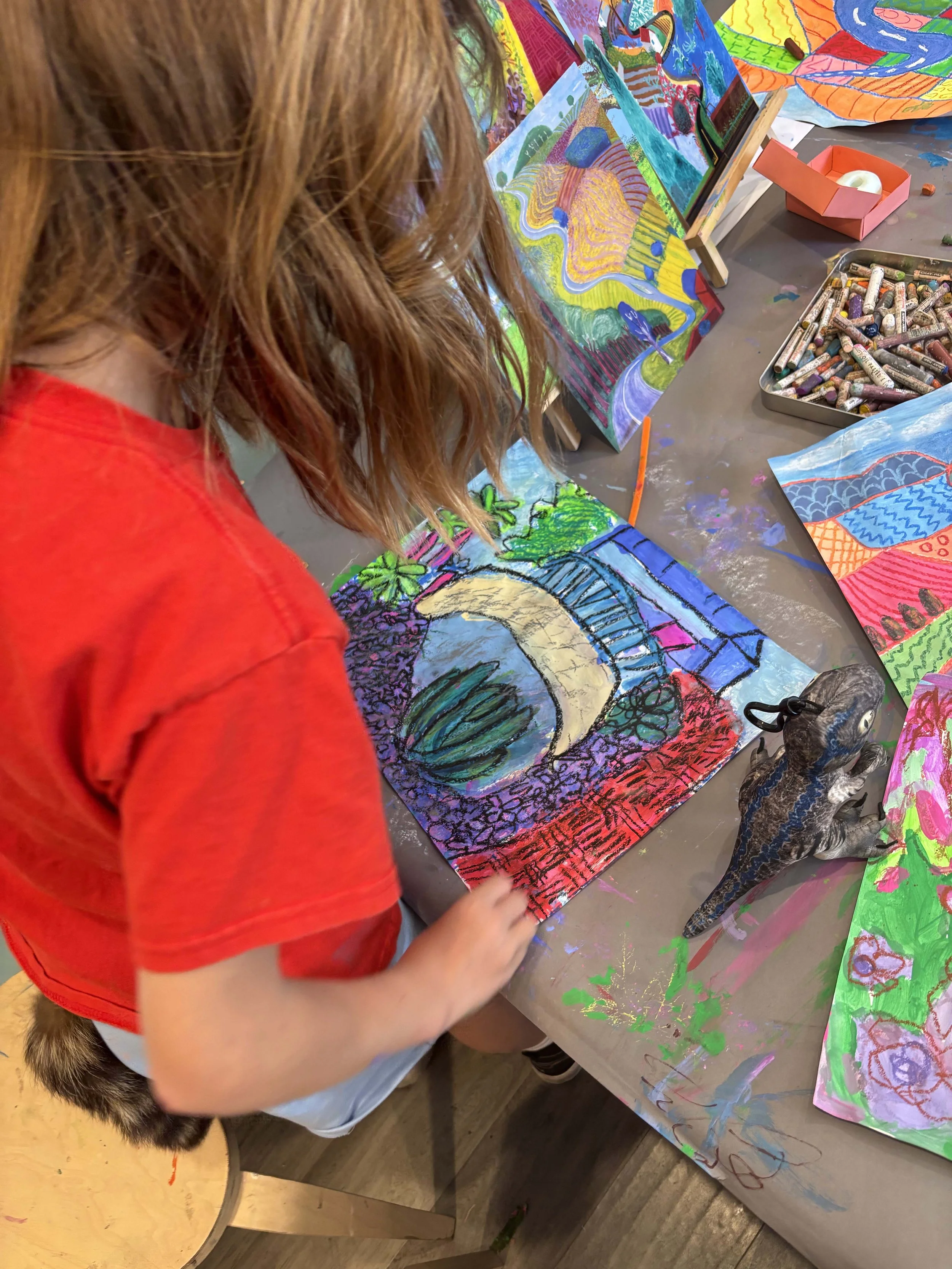 Child with red hair and a red shirt drawing on a colorful piece of art with various patterns and images, surrounded by art supplies on the table.
