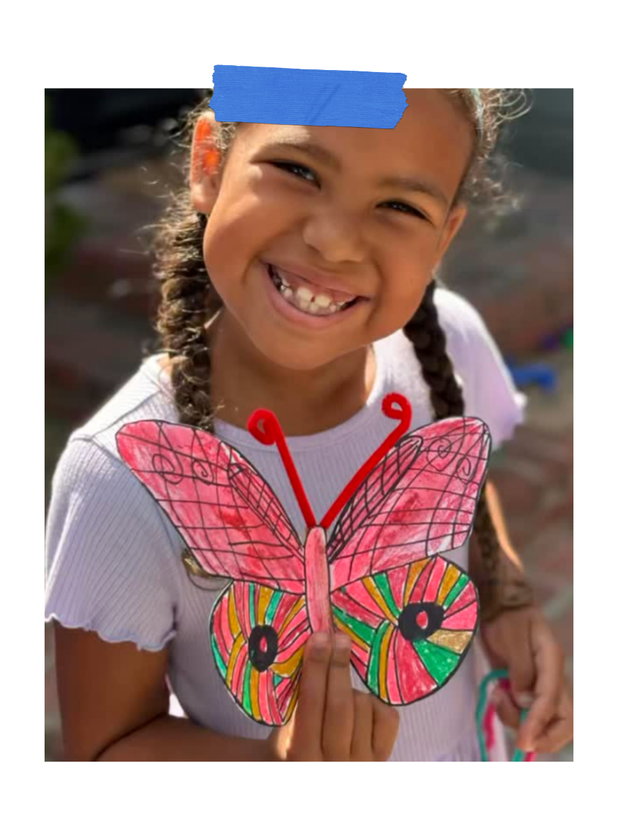 A young girl with braided hair is smiling and holding a colorful, hand-drawn butterfly craft. She is outdoors with sunlight illuminating her face.