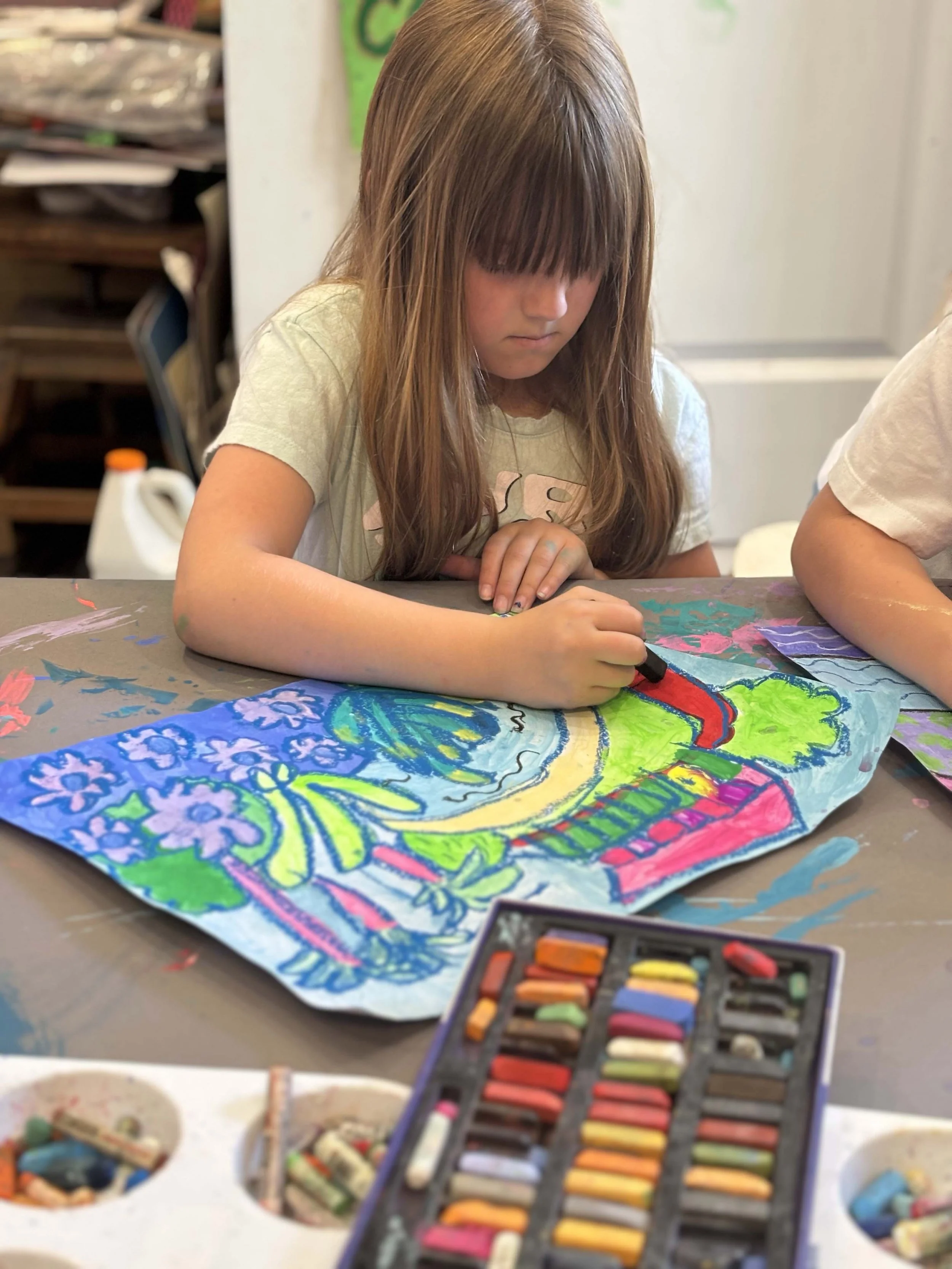 A young girl with long brown hair and bangs is focused on drawing and coloring on a large piece of paper with vibrant colors, using pastels or crayons, in an art classroom or studio.