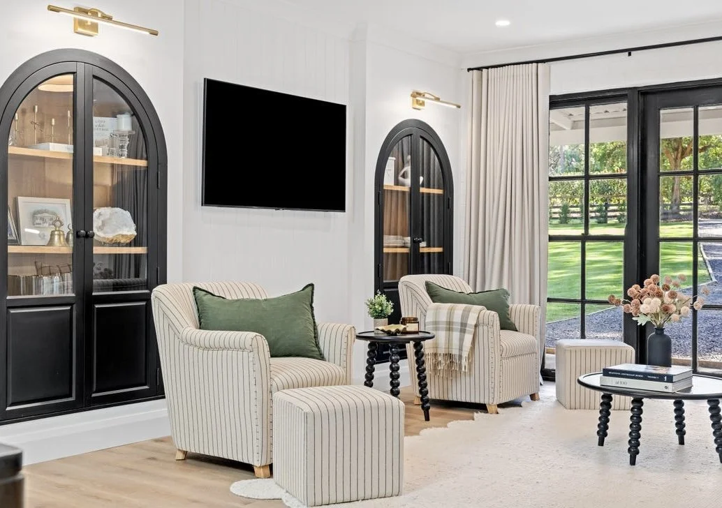 Living room with two striped armchairs with green pillows, side tables, a black coffee table with books and a vase of flowers, black glass-front cabinets, a wall-mounted flat-screen TV, sliding glass doors with white curtains, and a view of a yard with trees outside.