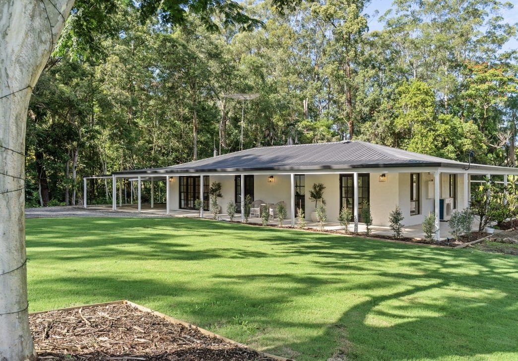Single-story house with white exterior, black-framed windows, surrounded by a well-maintained green lawn and trees in the background, with a porch running along the front.