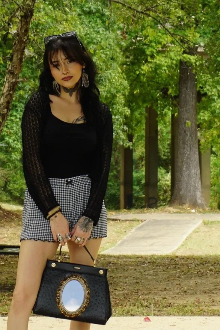 A woman standing outdoors in a park, wearing a black long-sleeve top with lace sleeves, checkered shorts, and holding a black handbag with an ornate oval mirror design. She has dark hair, makeup, and is accessorized with glasses on her head, earrings, necklaces, and rings.