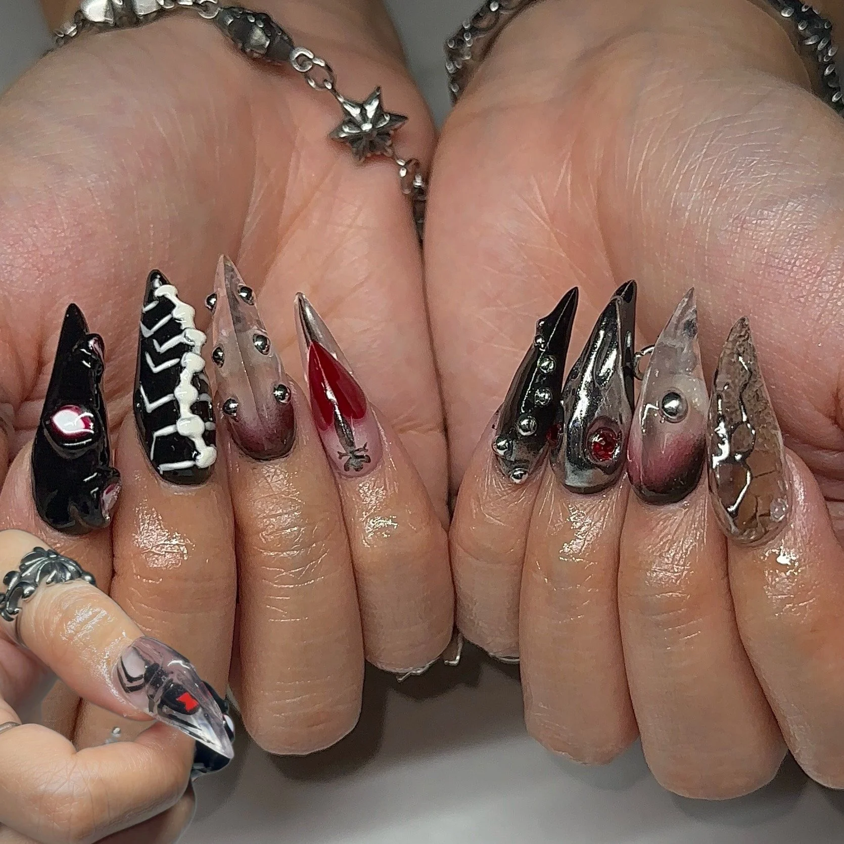 Close-up of hands with elaborate, decorated acrylic nails featuring black, white, red, and whodden designs, and wearing silver rings and bracelets.