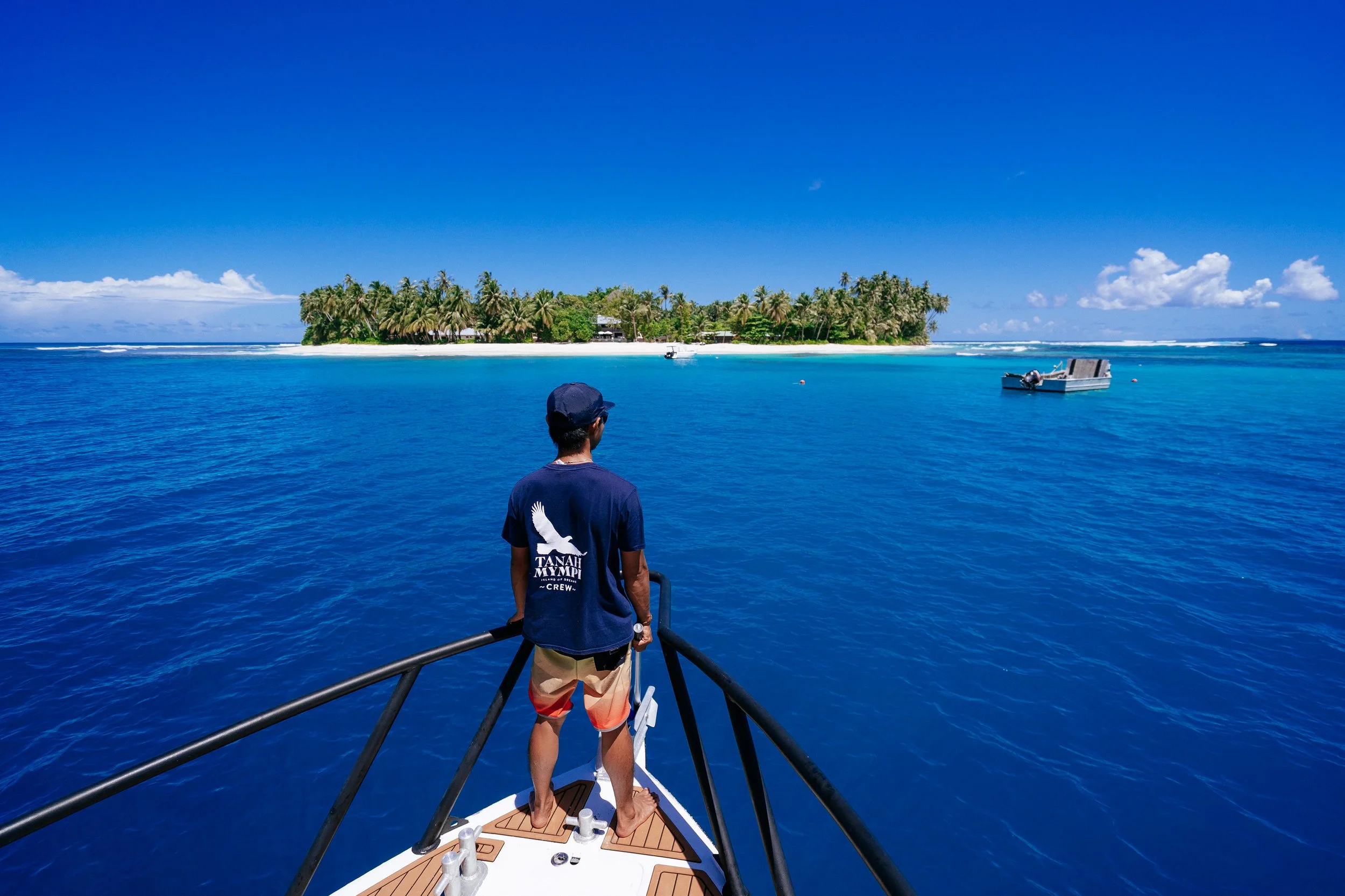 A Tanah Mympi crew member stands at the bow of a boat, looking out over the reef toward a nearby island.