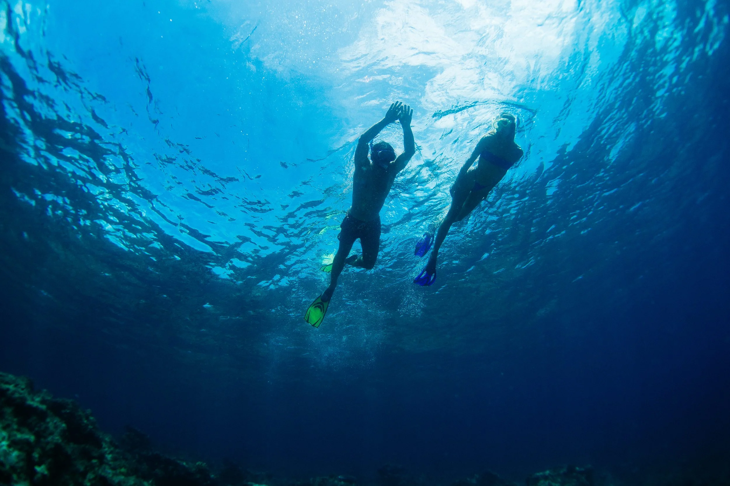 Two snorkelers swim through clear water off Tanah Mympi, sunlight streaming down from the surface above.