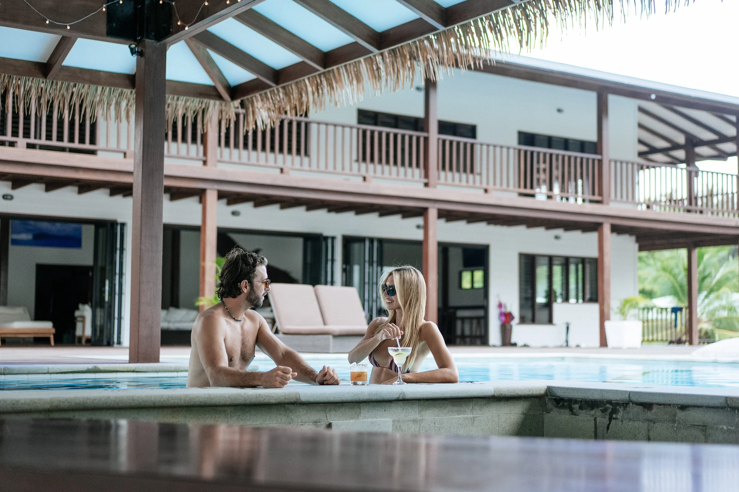 A couple relaxes at the edge of an infinity pool at Tanah Mympi, drinks in hand, with the jungle lined building behind them.