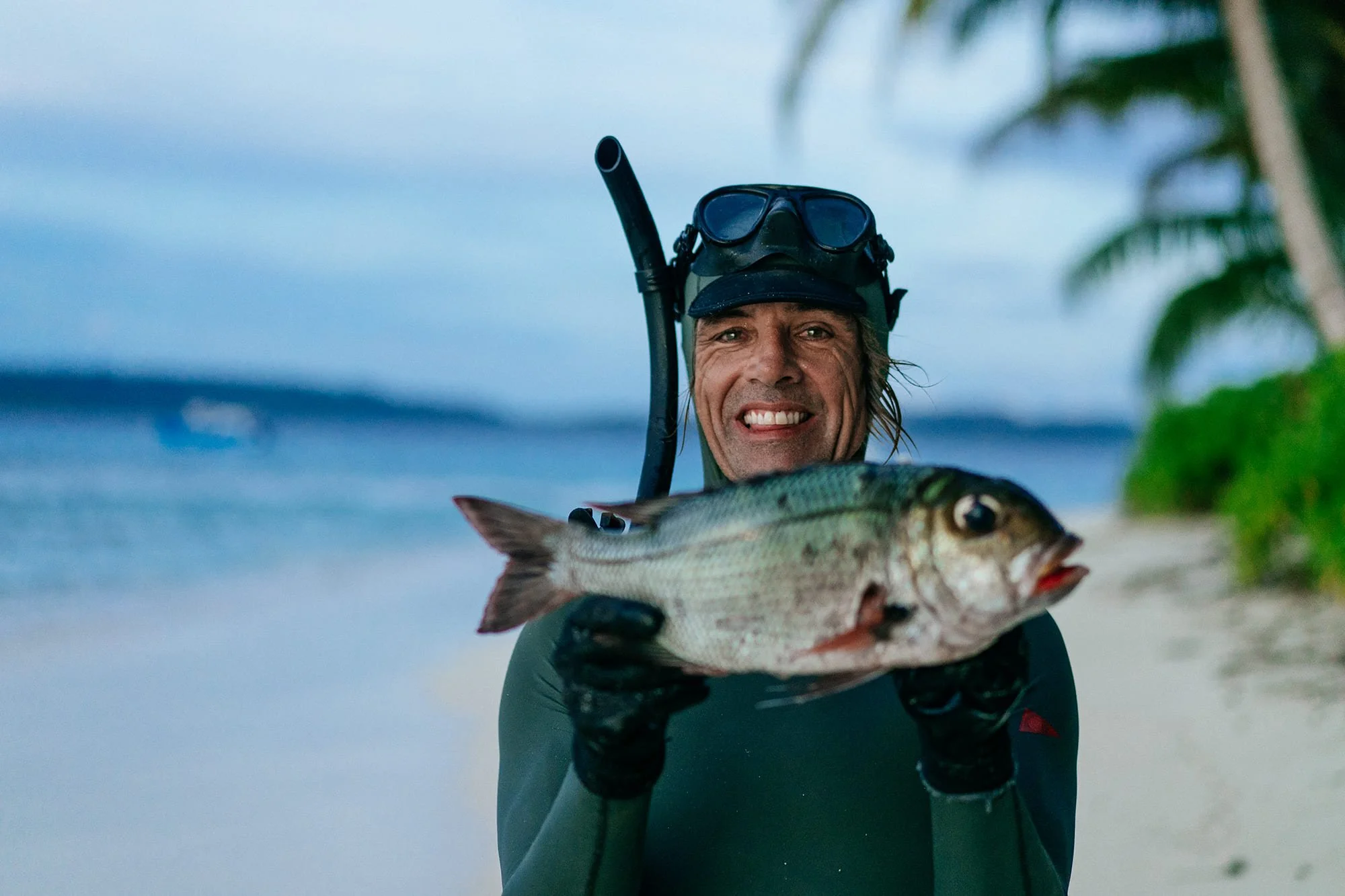 A smiling snorkeler holds up a freshly caught fish on the beach at Tanah Mympi, mask and snorkel still on.
