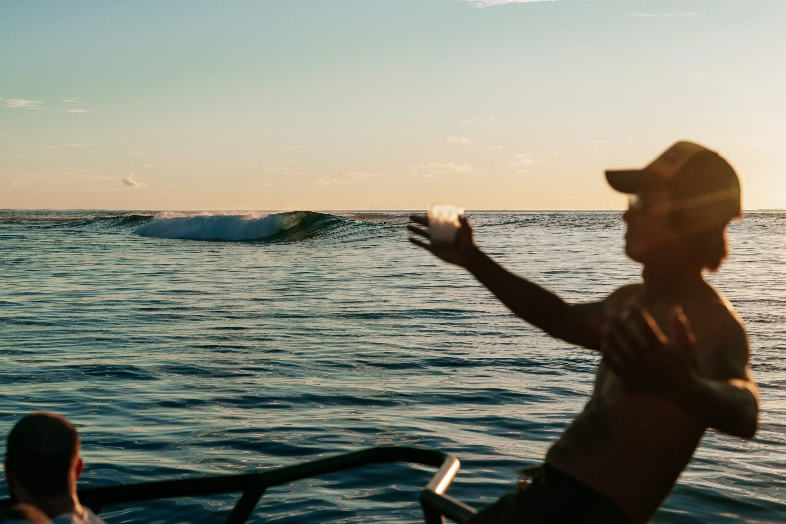 A guest silhouetted against a golden sunset, reaching up from the deck of a boat on the water near Tanah Mympi.