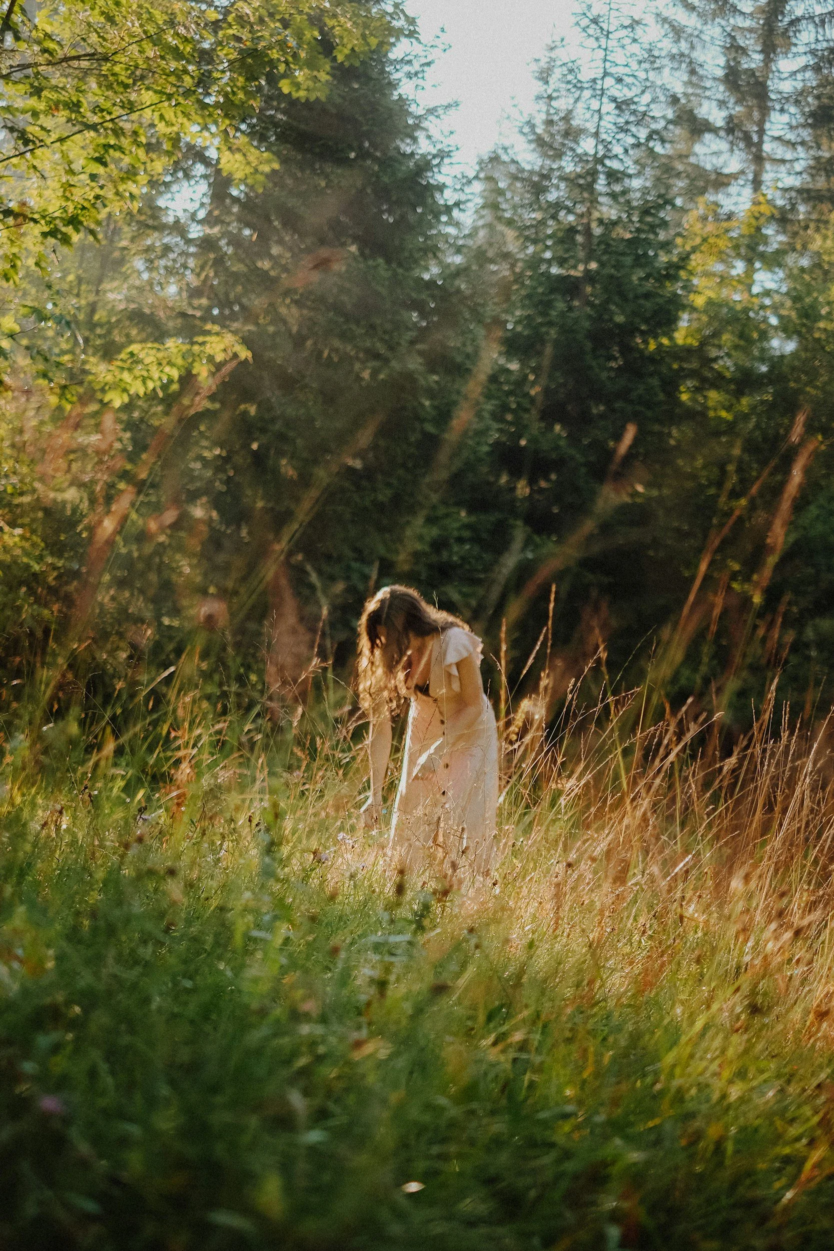 A person with long hair wearing a light-colored dress is in a grassy field surrounded by tall grass and trees, with sunlight streaming through the foliage.