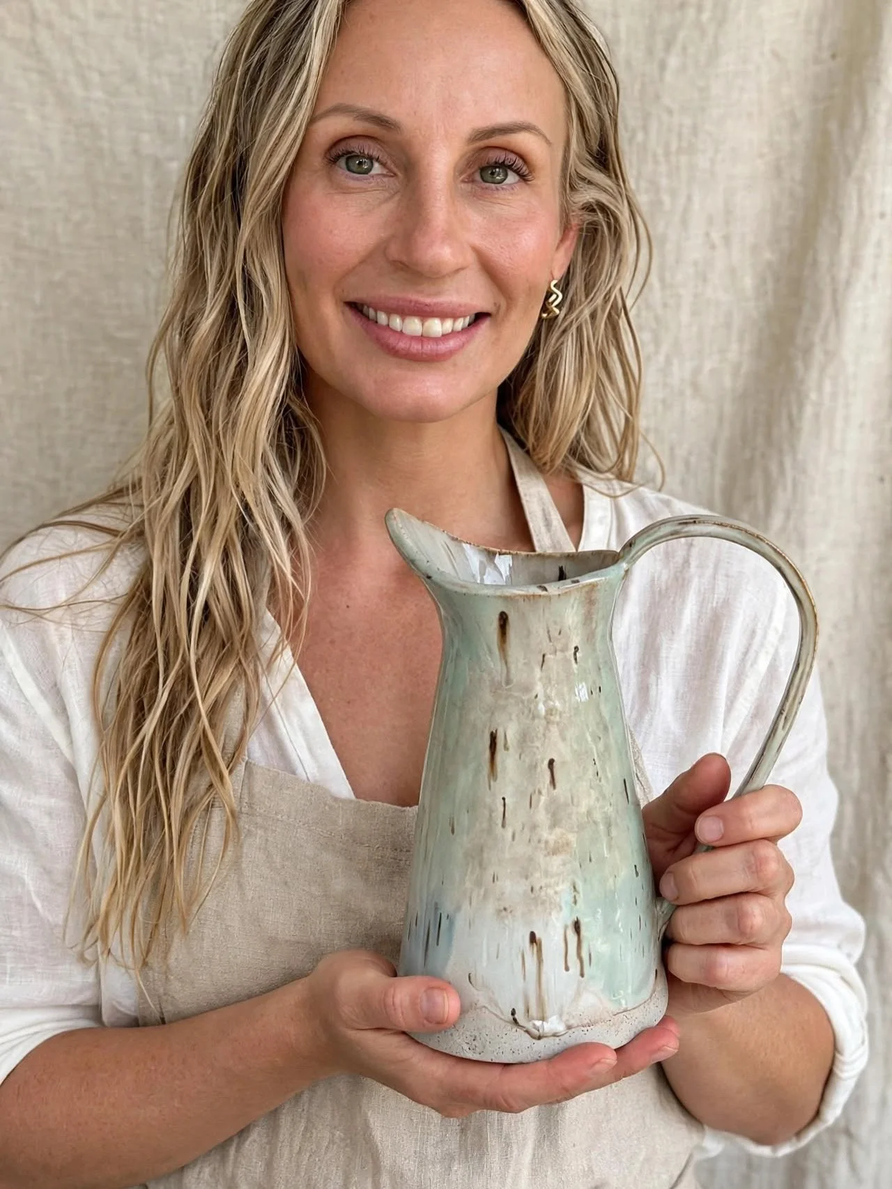 A woman with blonde, curly hair and earrings smiling while holding a ceramic pitcher.