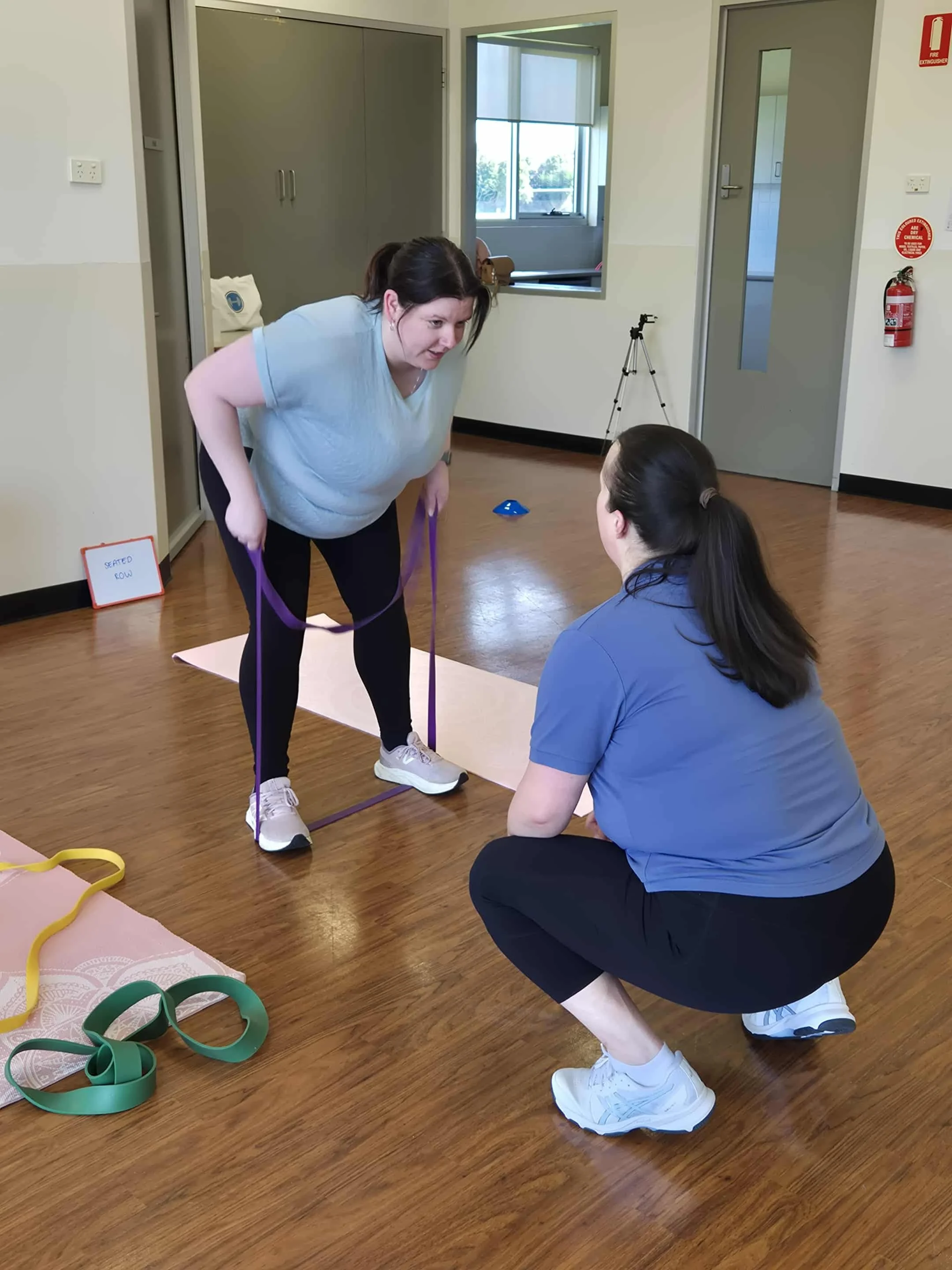 is assisting a woman in a blue shirt and black pants with a stretching or yoga exercise using a purple resistance band in a room with hardwood floors. There are colorful resistance bands on the floor nearby, and a small sign that reads 'Seated Row' in the background.