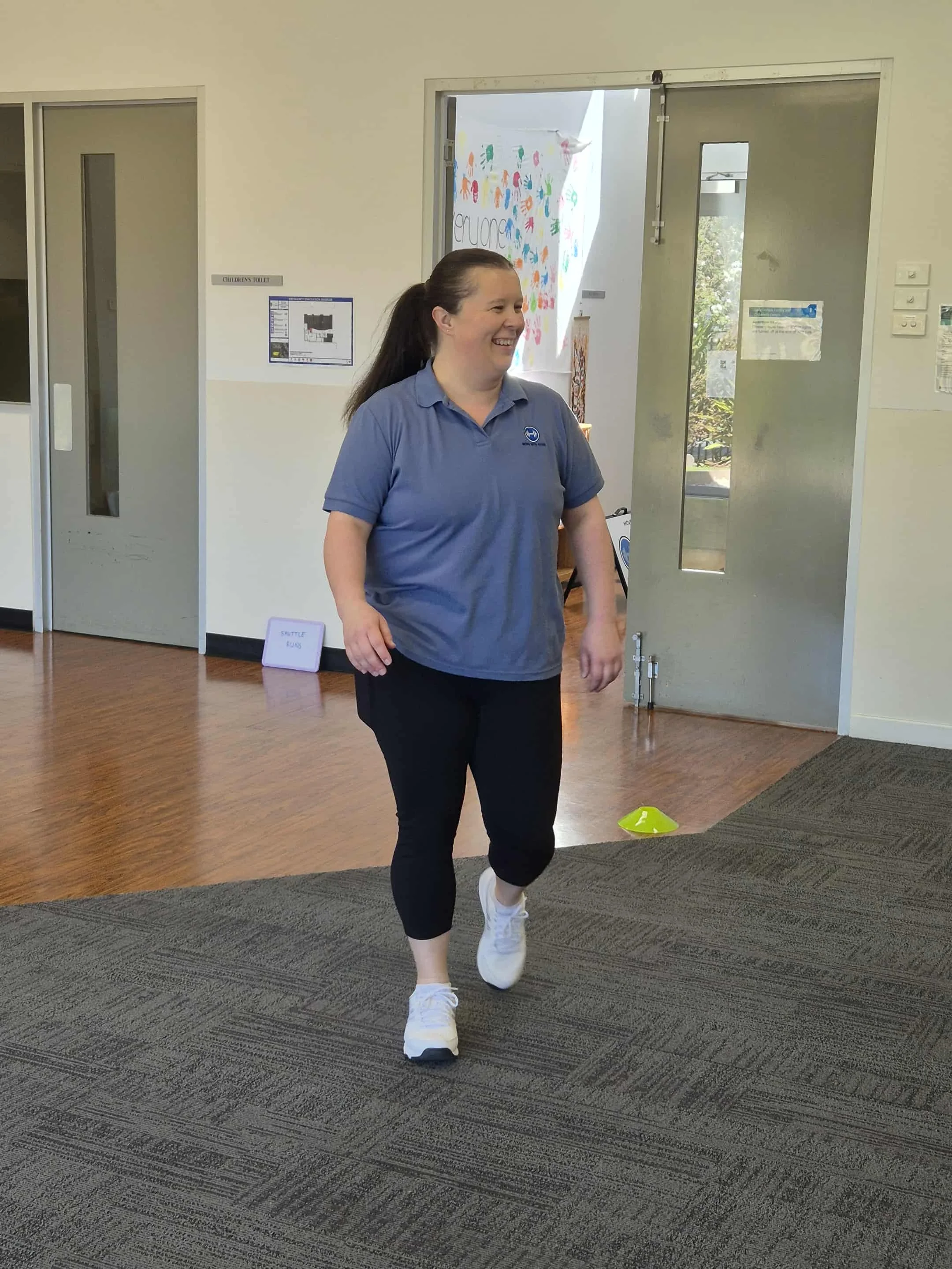 A woman walking indoors with a smile, wearing a blue polo shirt, black pants, and white sneakers, near a door with a colorful wall hanging in the background.