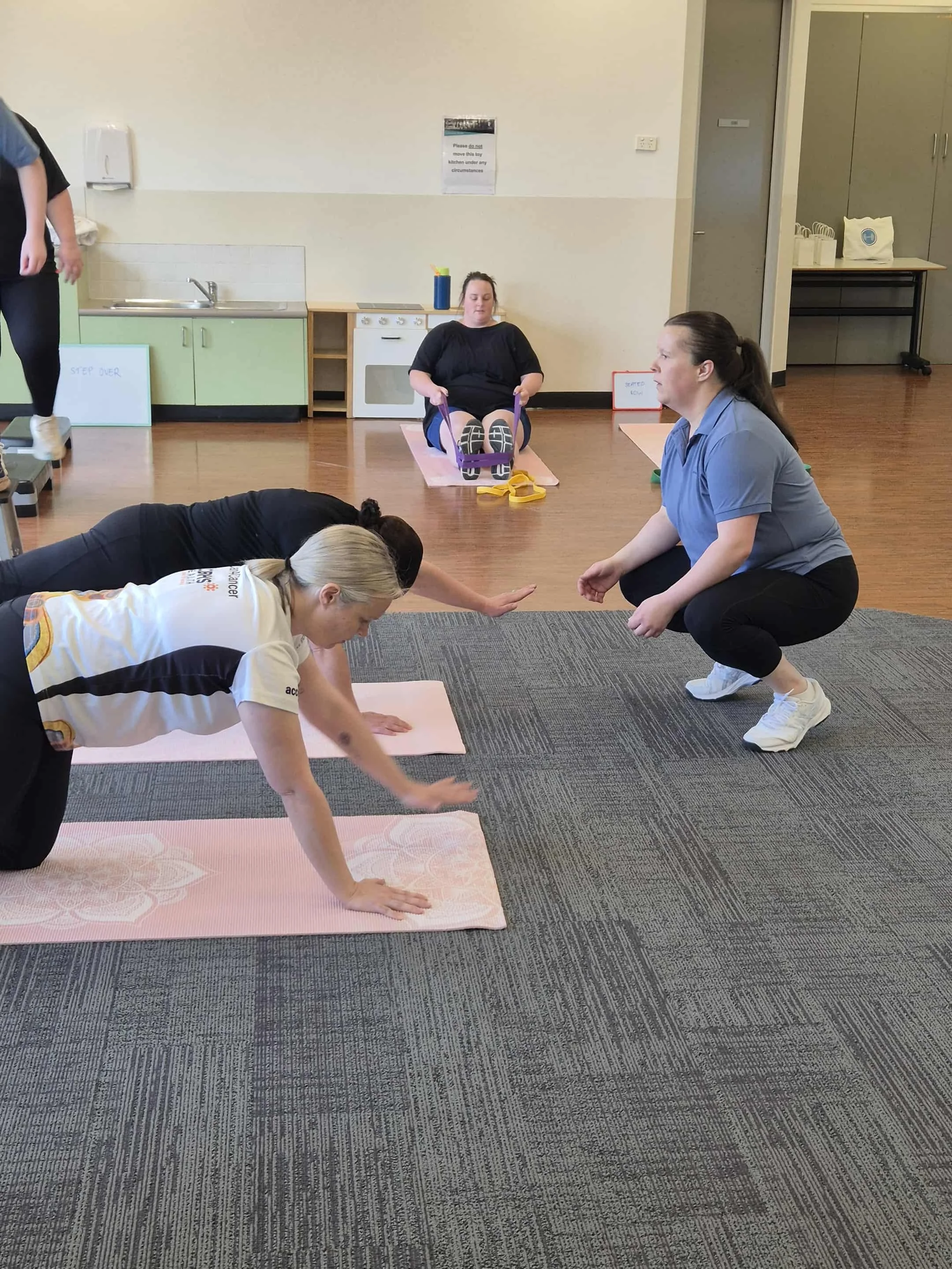 Group of people participating in a yoga or exercise class in a room with wooden floors, some on yoga mats, with a sink and cabinets in the background.