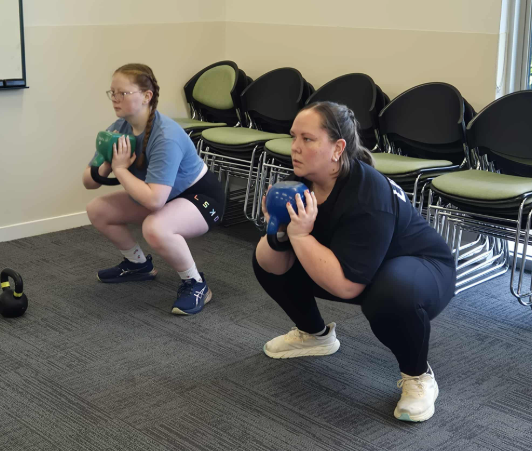Two women performing squat exercises with kettlebells in a room with stacked chairs and a carpeted floor.