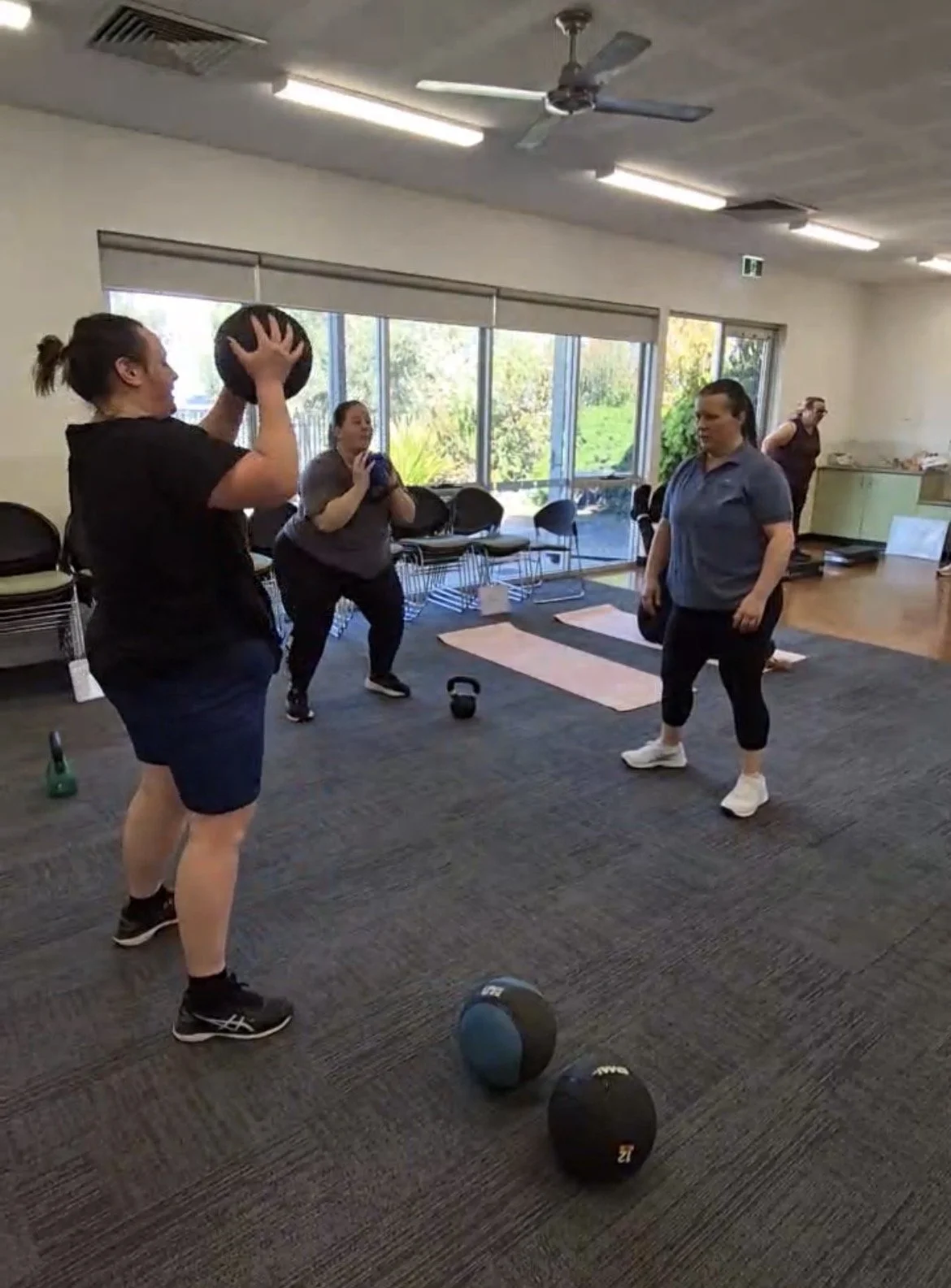 Four women participating in a fitness class with kettlebells and medicine balls in a room with large windows, chairs, and exercise mats.
