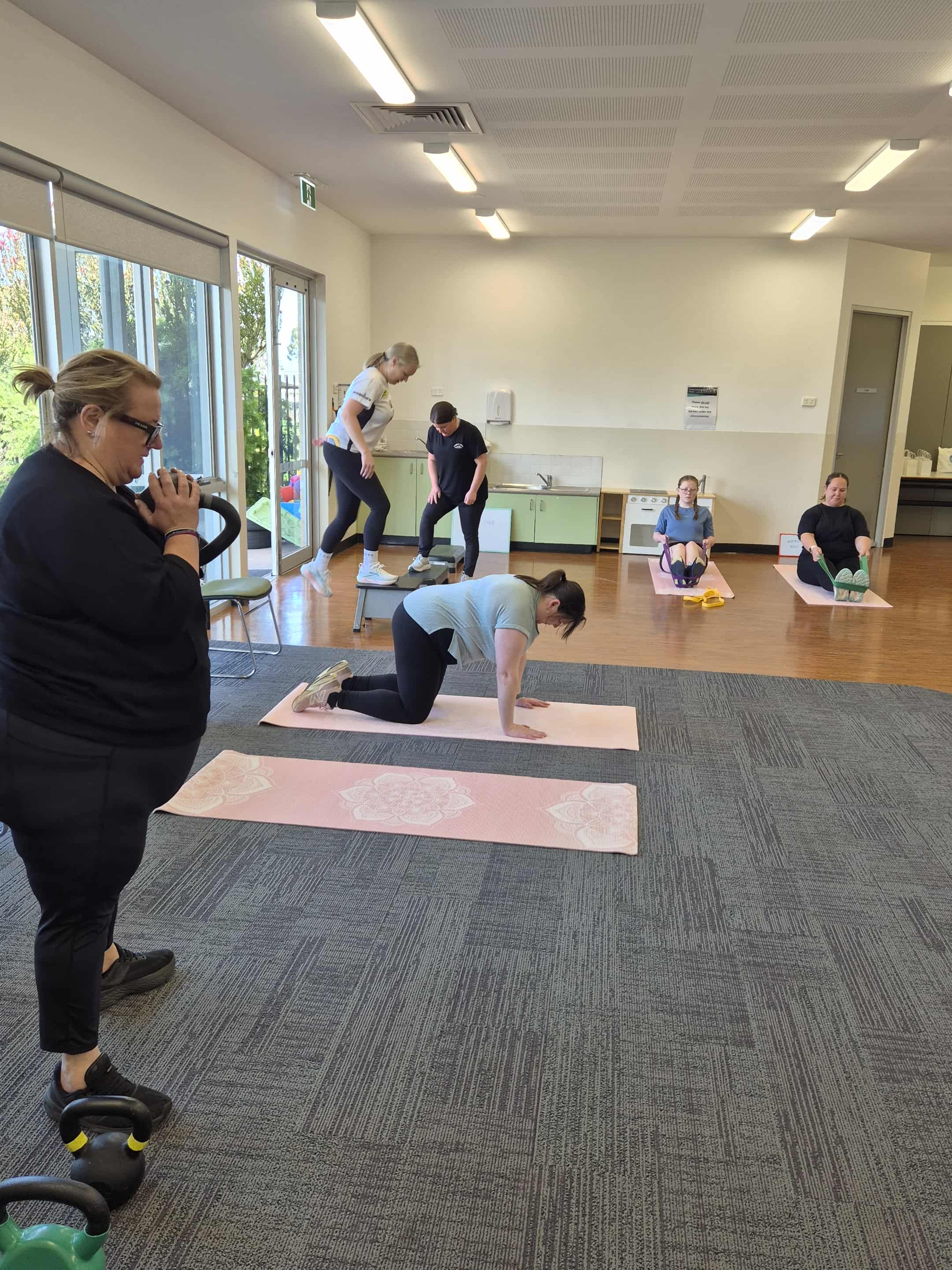 Group of women participating in a fitness or exercise class in a room. One woman is doing a plank on a pink yoga mat, others are on yoga mats or standing, with some observing or coaching.