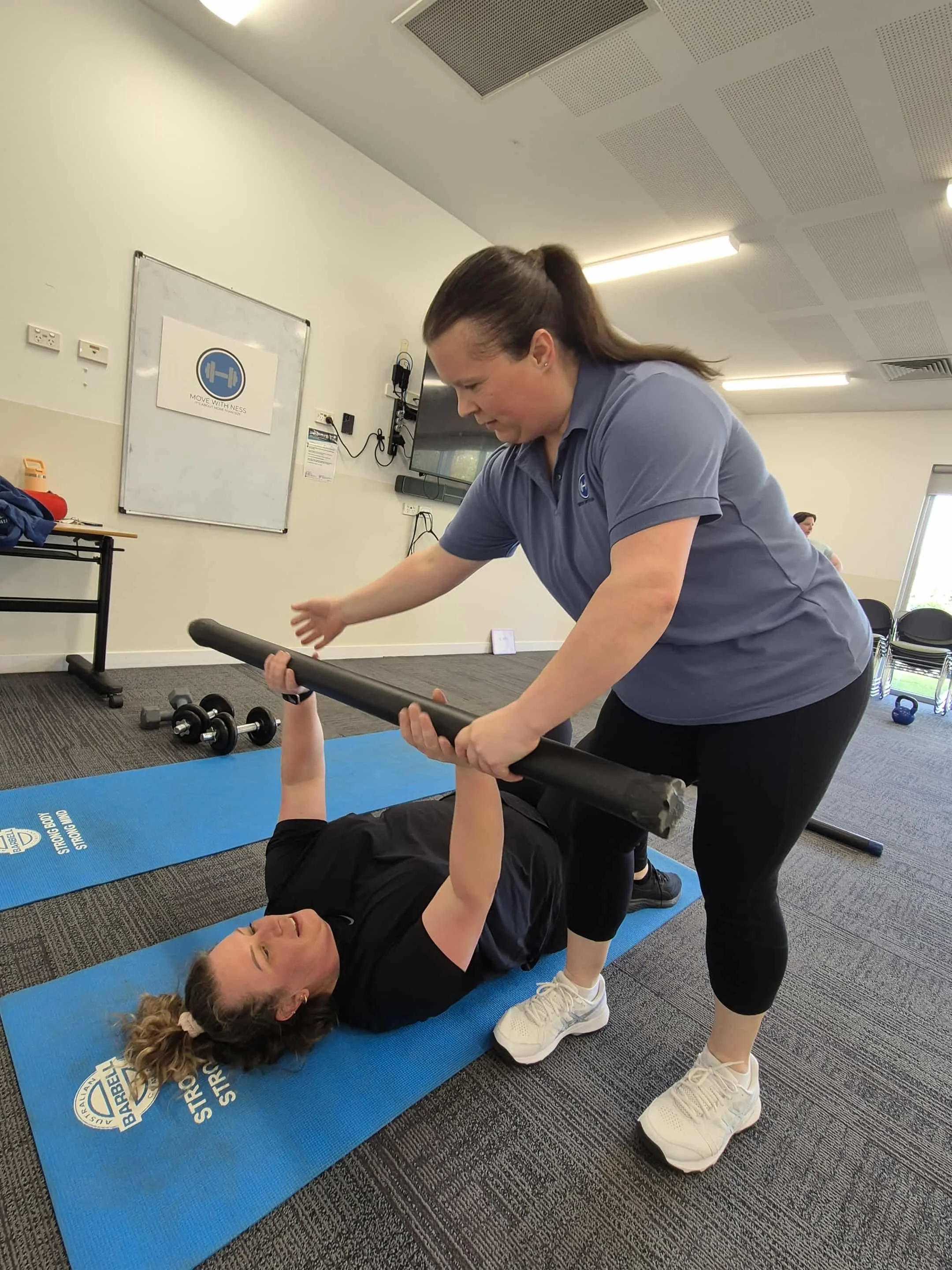 Ness assisting a woman lying on a blue exercise mat by holding a revo bar. The woman on the mat is lifting the bar with both hands while the helper supports from below. The setting appears to be a gym or fitness studio.