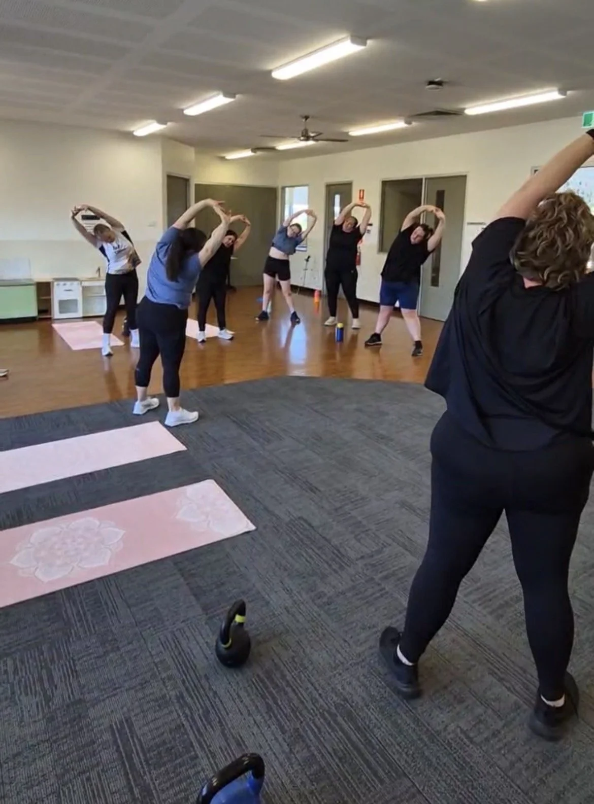 A group of people participating in a stretching or yoga class, standing with arms over their heads, in a room with wooden floors, pink exercise mats, and several ceiling lights.