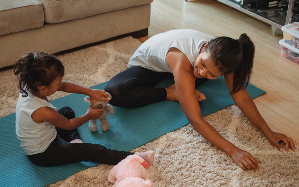 A woman and a young girl practicing yoga on a mat in a living room, with the girl playing with a stuffed animal.