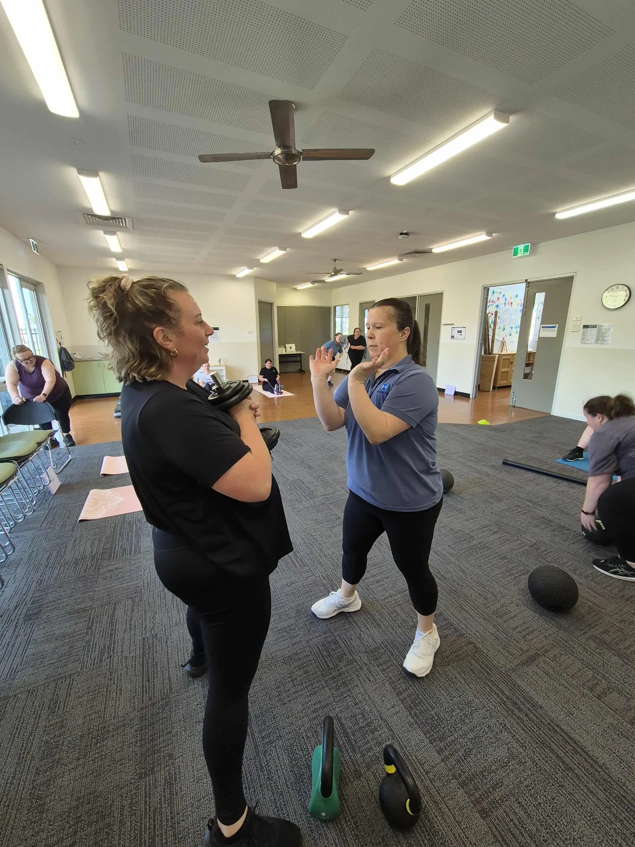 Two women are engaged in a fitness or exercise class inside a bright room with several other people in the background, with fitness equipment like kettlebells on the floor nearby. One woman is holding dumbbells, and the other is demonstrating an exercise. The room has chairs, a clock, and large windows letting in natural light.