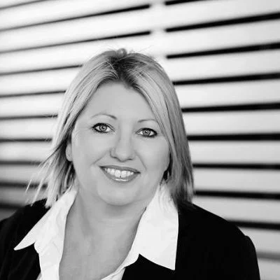 Black and white photo of a woman smiling, with shoulder-length hair, wearing a white shirt and dark blazer, standing in front of a striped background.