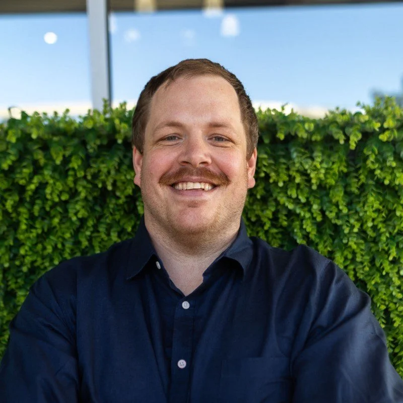 A smiling man with short light brown hair, wearing a navy blue button-up shirt, sitting in front of green bushes in an outdoor setting.