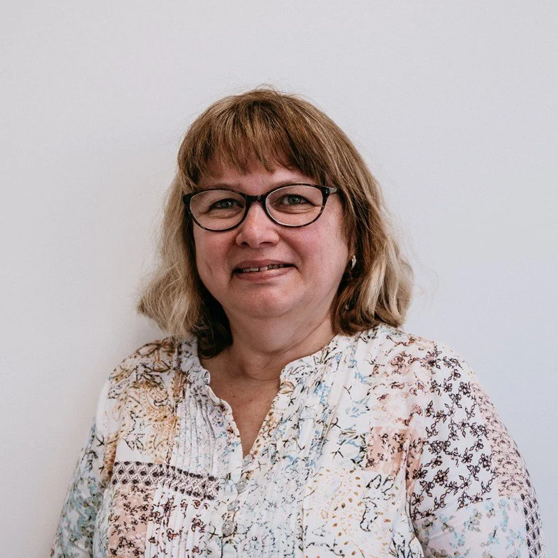 A middle-aged woman with blonde, wavy hair and black glasses smiling at the camera, wearing a patterned blouse, standing against a plain white wall.