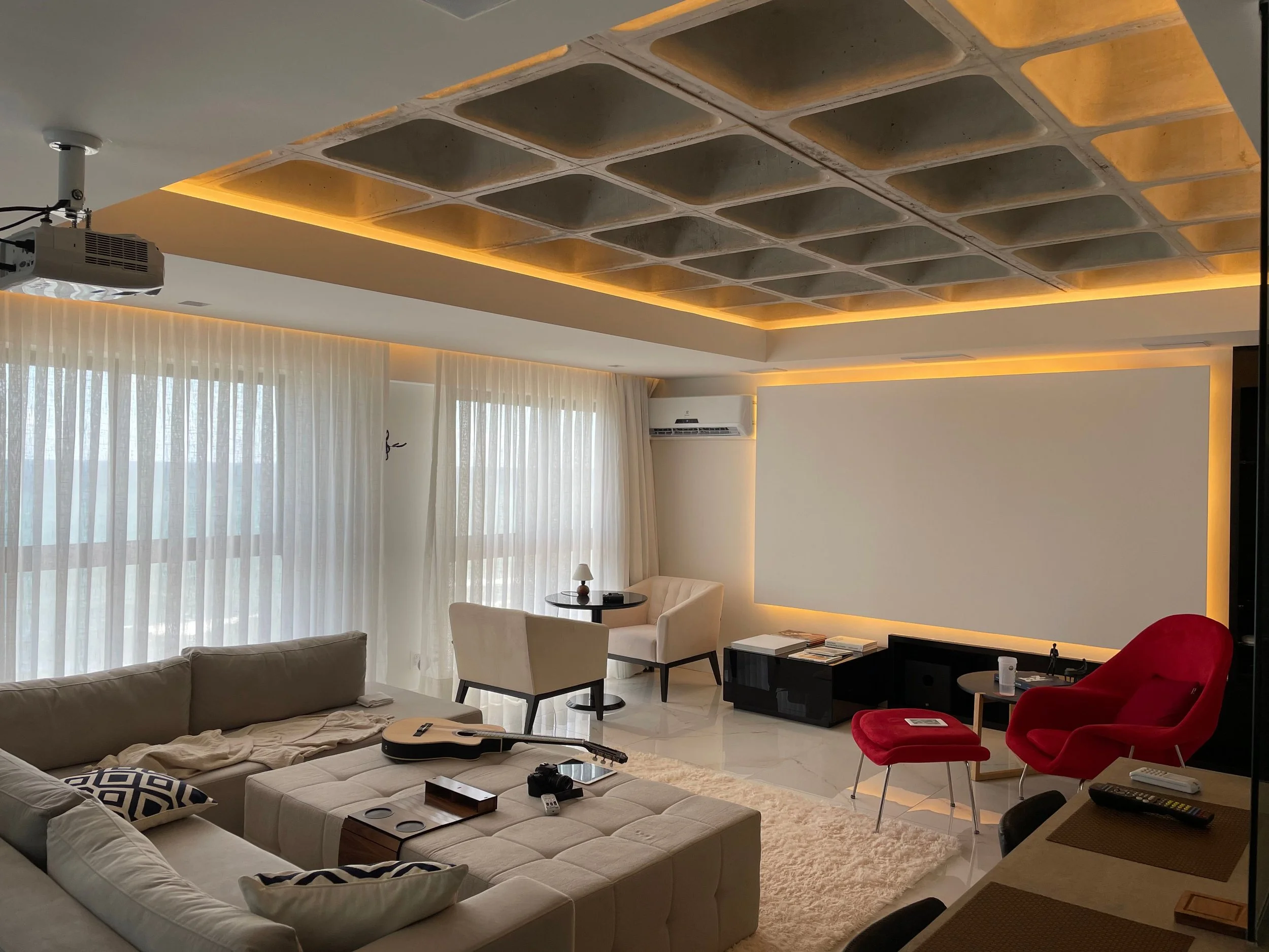 Modern living room with beige sectional sofa, red armchairs, round coffee table, and ceiling with built-in lighting. Large window with white curtains, wall-mounted air conditioner, and a projector mounted on ceiling.