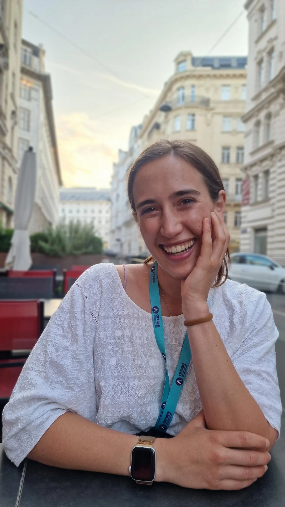 A young woman with brown hair smiling, seated outdoors at a table in an urban area, wearing a white patterned top, a smartwatch, and a lanyard. The background shows European-style buildings and a street.