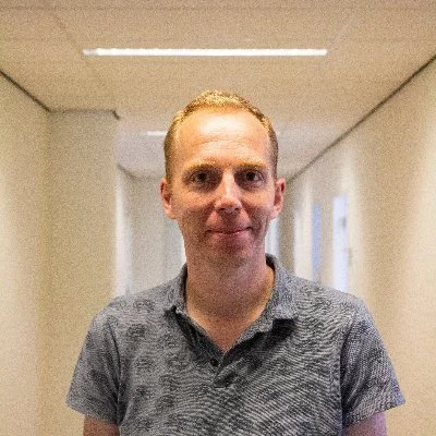 Close up of Jeroen with short blonde hair and wearing a gray short-sleeve polo shirt standing in a hallway with white walls and ceiling lights.
