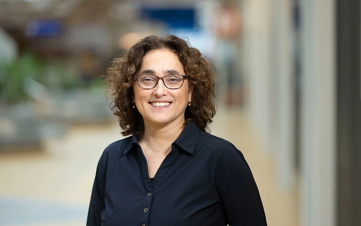 Close up of Gönül with curly brown hair, glasses, and a dark blue button-up shirt smiling in a blurred indoor setting.