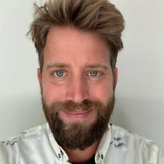 Close-up of a Jasper with light brown hair, a full beard, and blue eyes, smiling at the camera against a plain light-colored background.