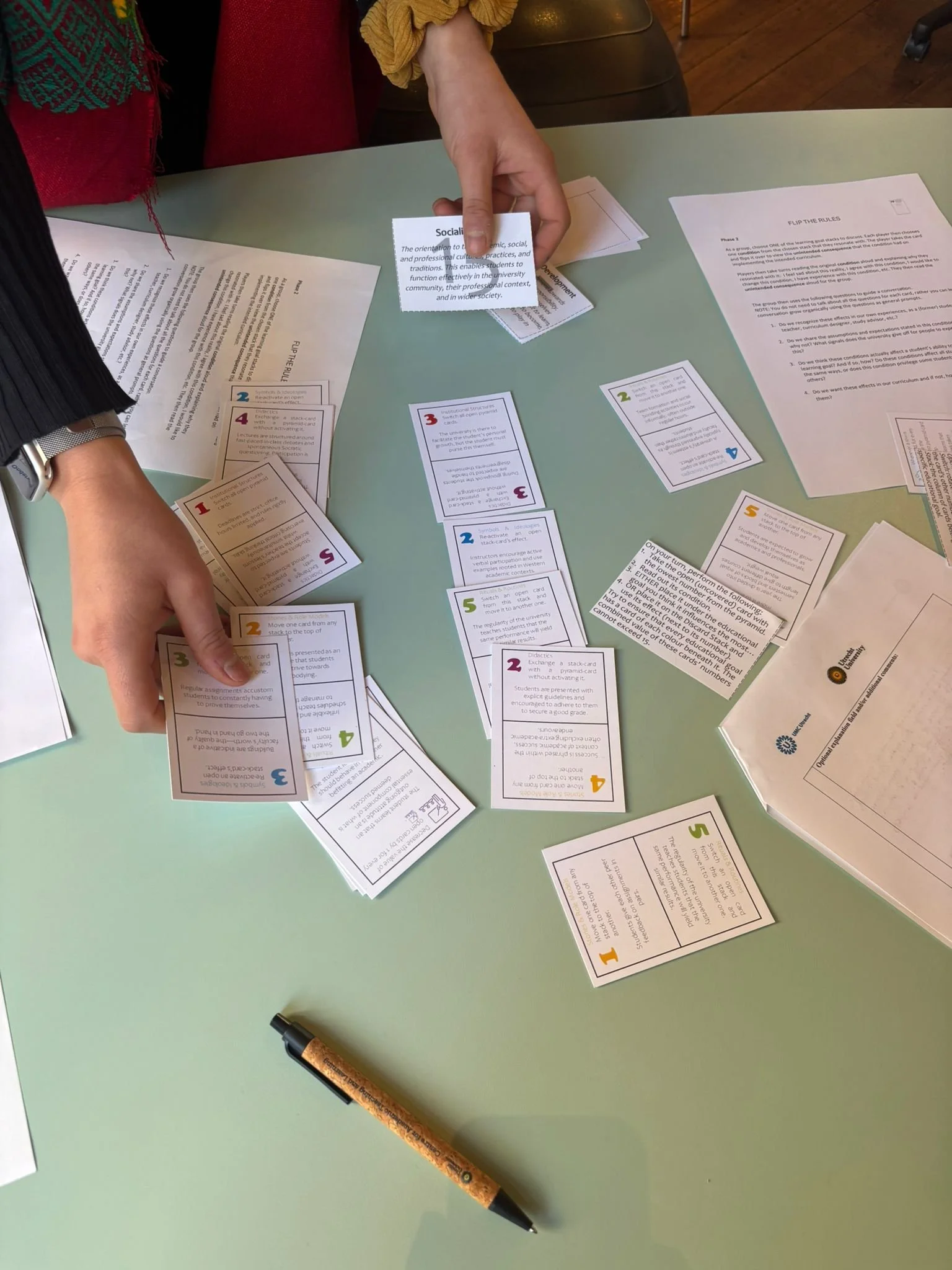 A person is sitting at a light green table, holding a small stack of cards with one card partly pulled out, and there are other cards spread across the table. The cards contain text and colorful numbers. A black and orange textured pen is on the table.