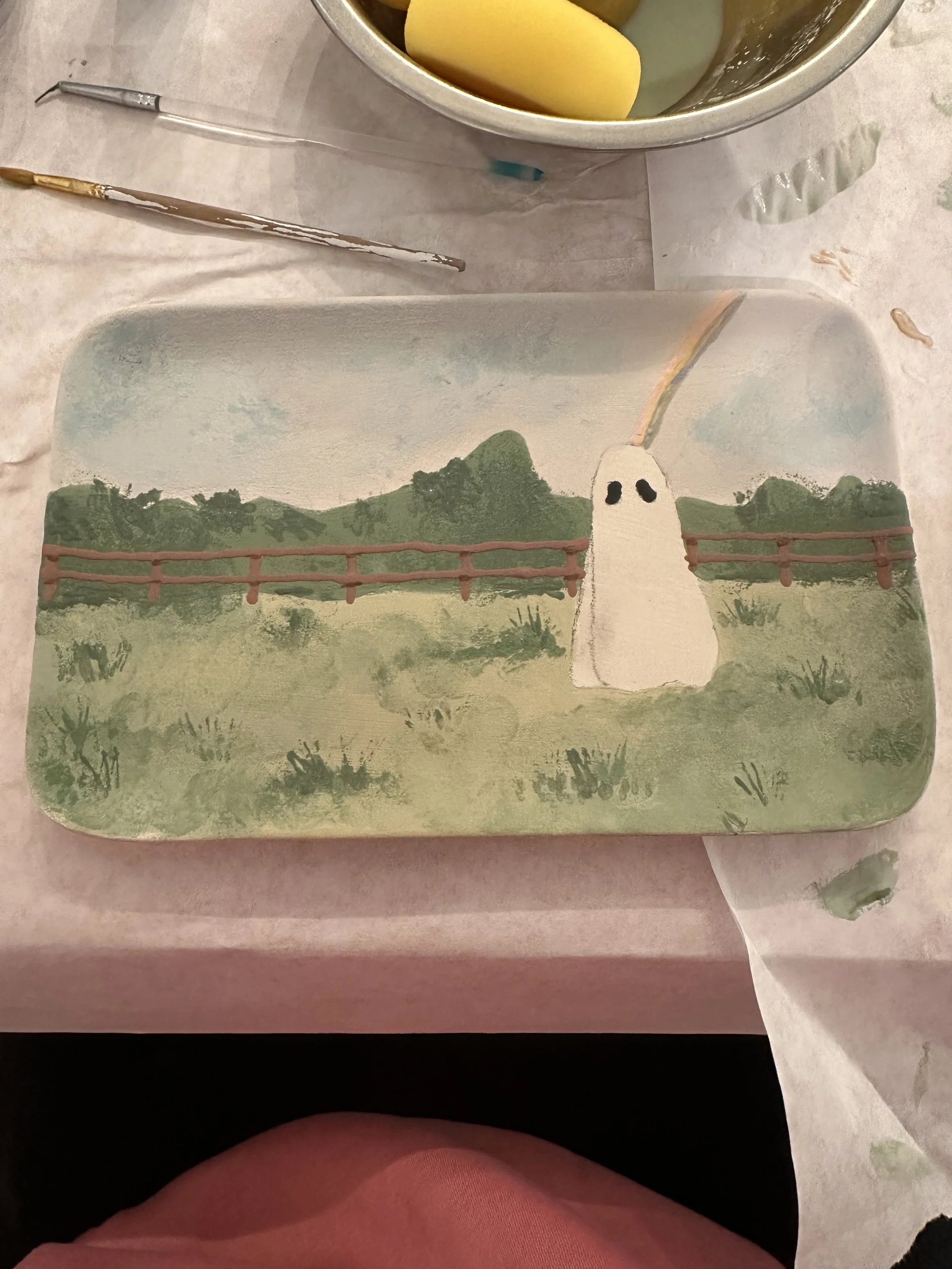 A painted landscape on a rectangular object featuring a tree with two black eyes, a bridge, mountains, and clouds, with some paint smudges and smears around.