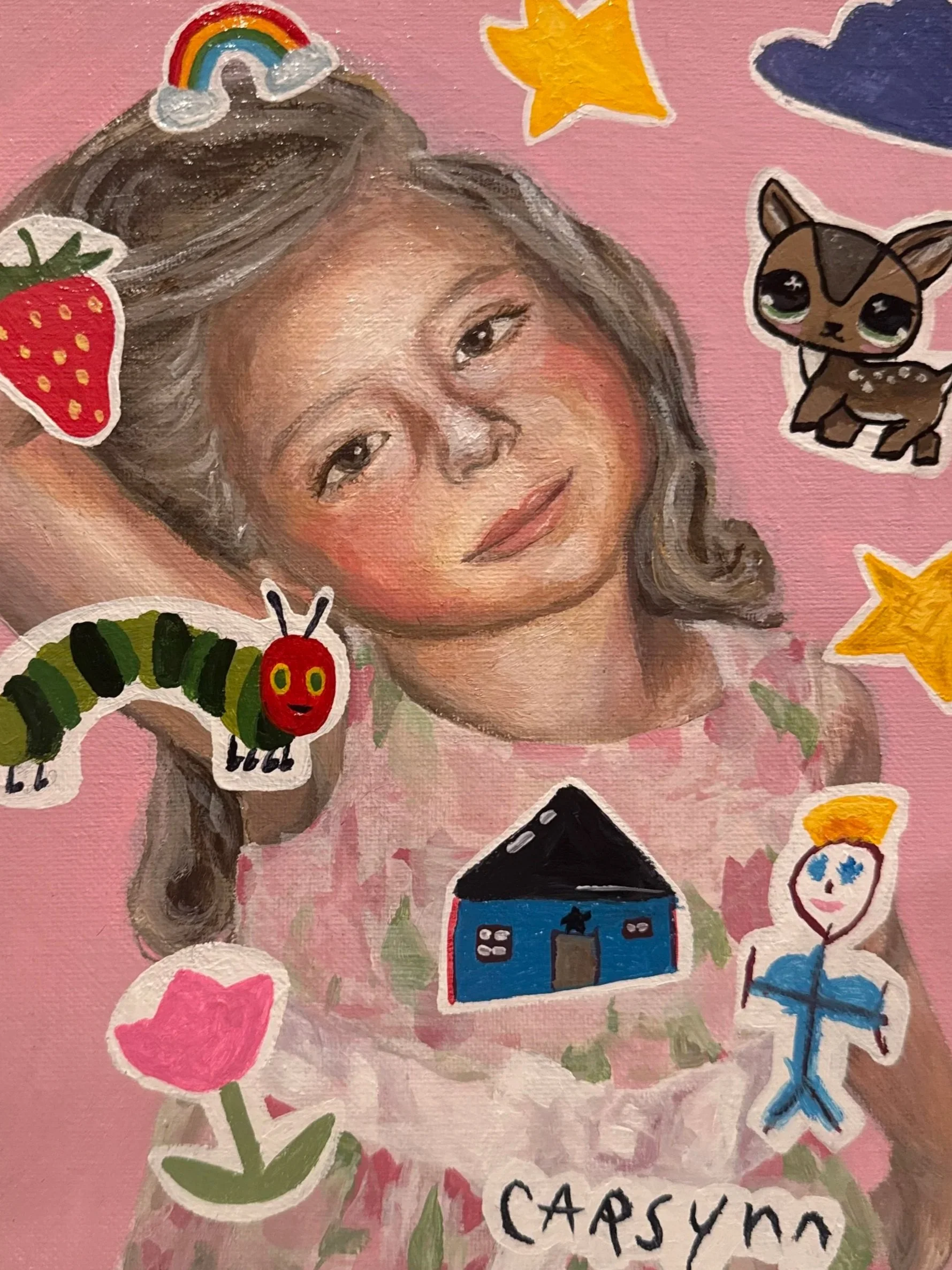 A painted portrait of a young girl with brown hair and eyes, set against a pink background. The painting is decorated with colorful stickers including a rainbow, star, dog, strawberry, caterpillar, pink heart, house, doll, and a blue starfish, along 