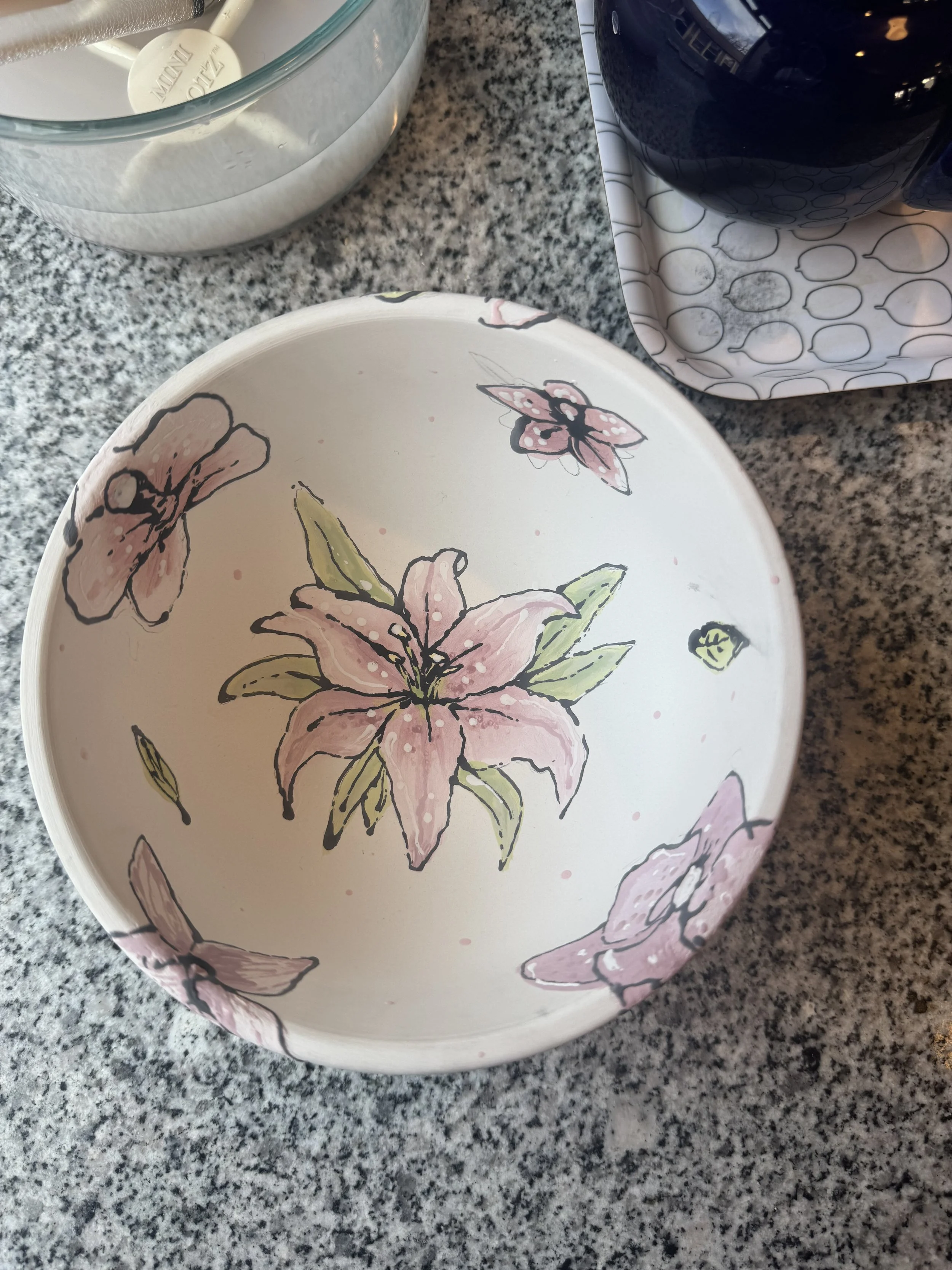 Close-up of a decorative ceramic plate with pink lilies and butterflies, placed on a speckled gray granite countertop, with a black object and paper napkin nearby.