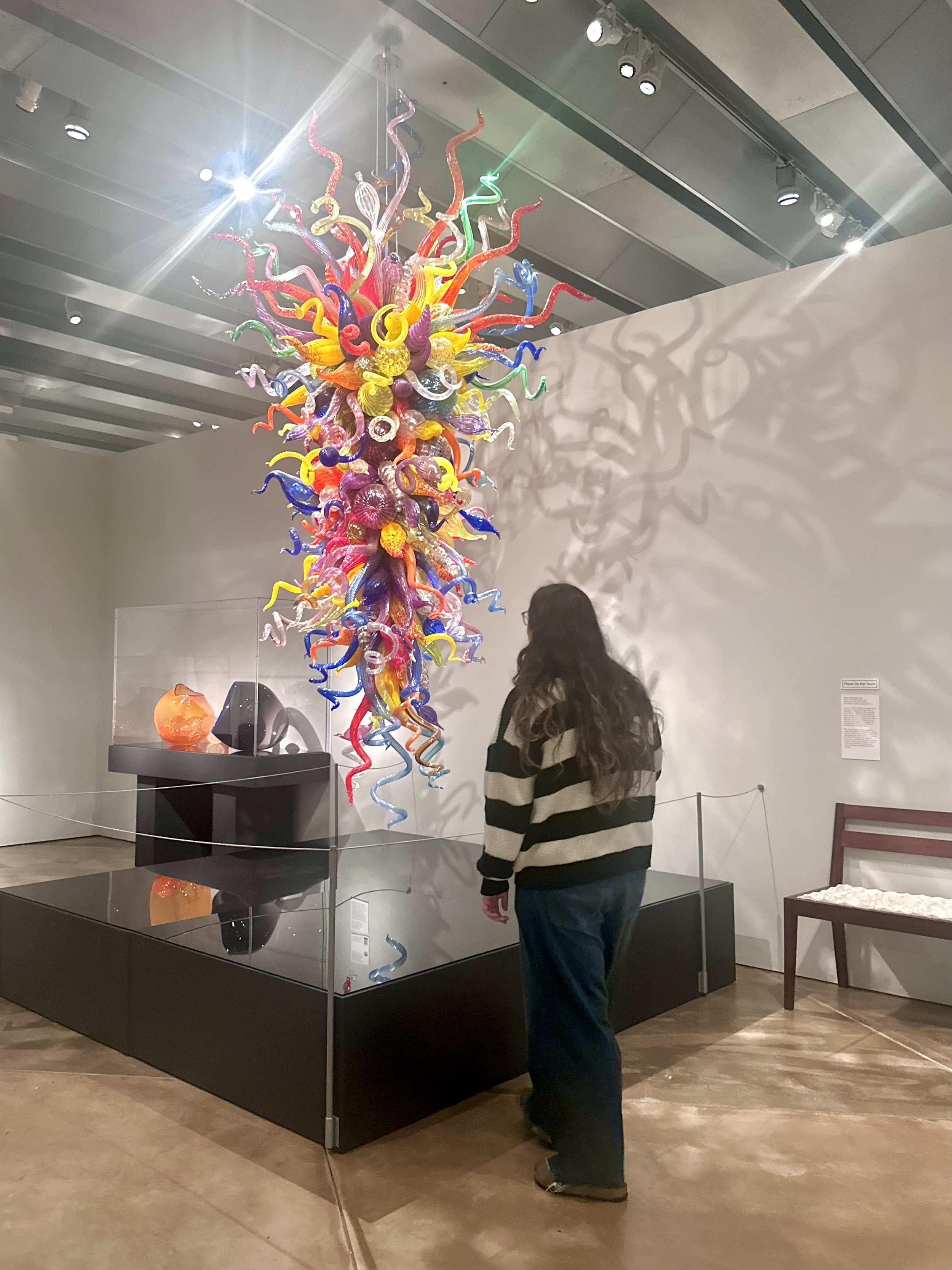 A woman with long dark hair wearing a black and tan striped sweater viewing a colorful glass sculpture of twisting, swirling glass pipes arranged in a large vertical cluster at a museum exhibit.