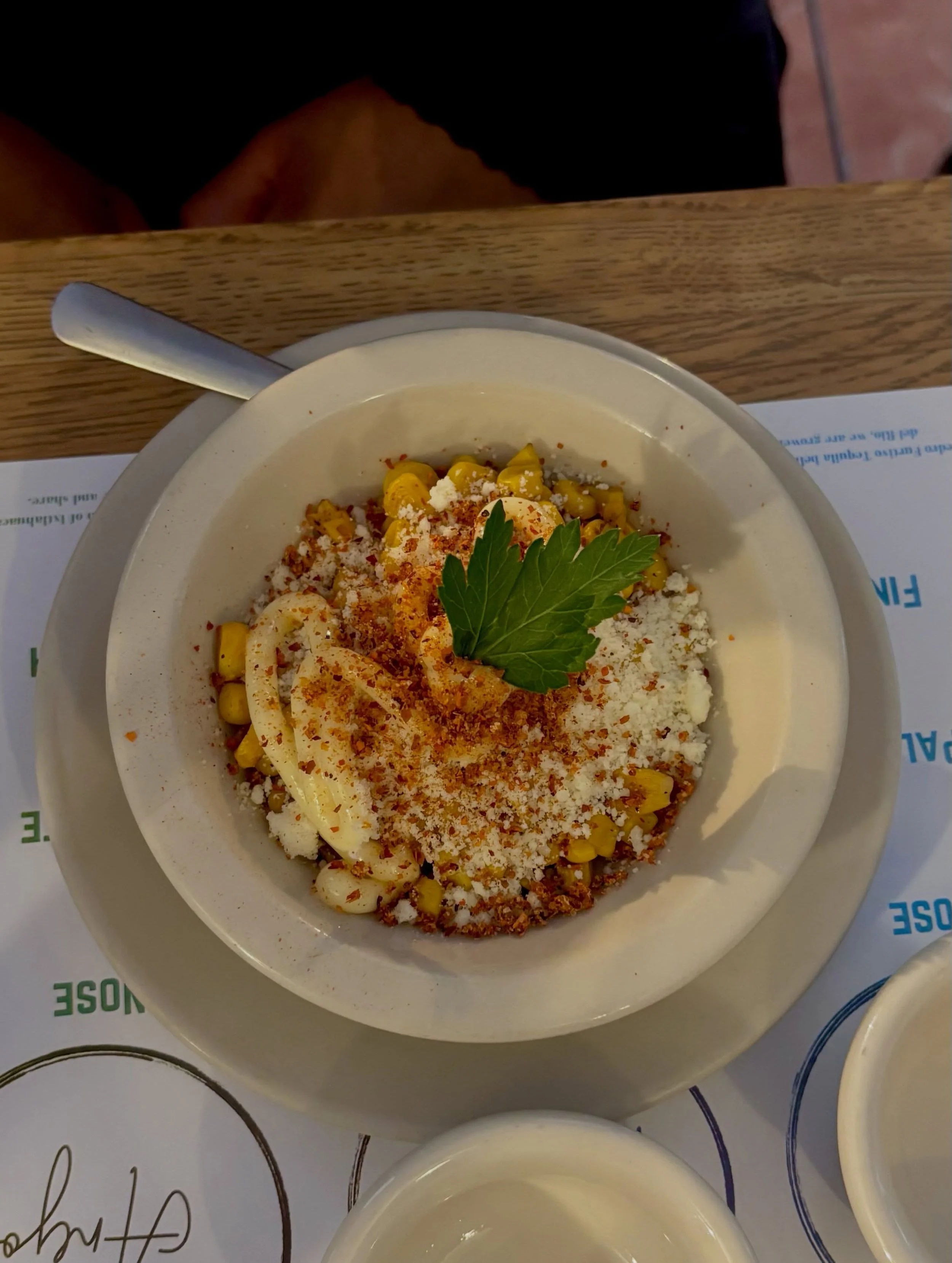 A bowl of Mexican corn soup garnished with a dollop of cream, chopped herbs, and sprinkled with grated cheese and chili powder, placed on a white plate on a table.