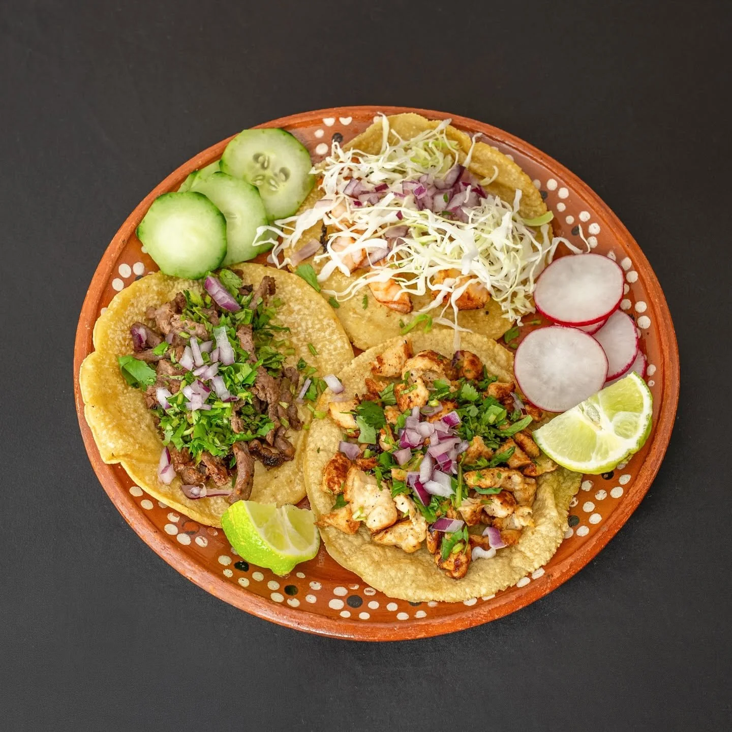 A round, orange terracotta plate filled with assorted Mexican dishes including tacos with chicken and beef, shredded cabbage, sliced cucumbers, radishes, lemon wedges, and lime slices on a black surface.