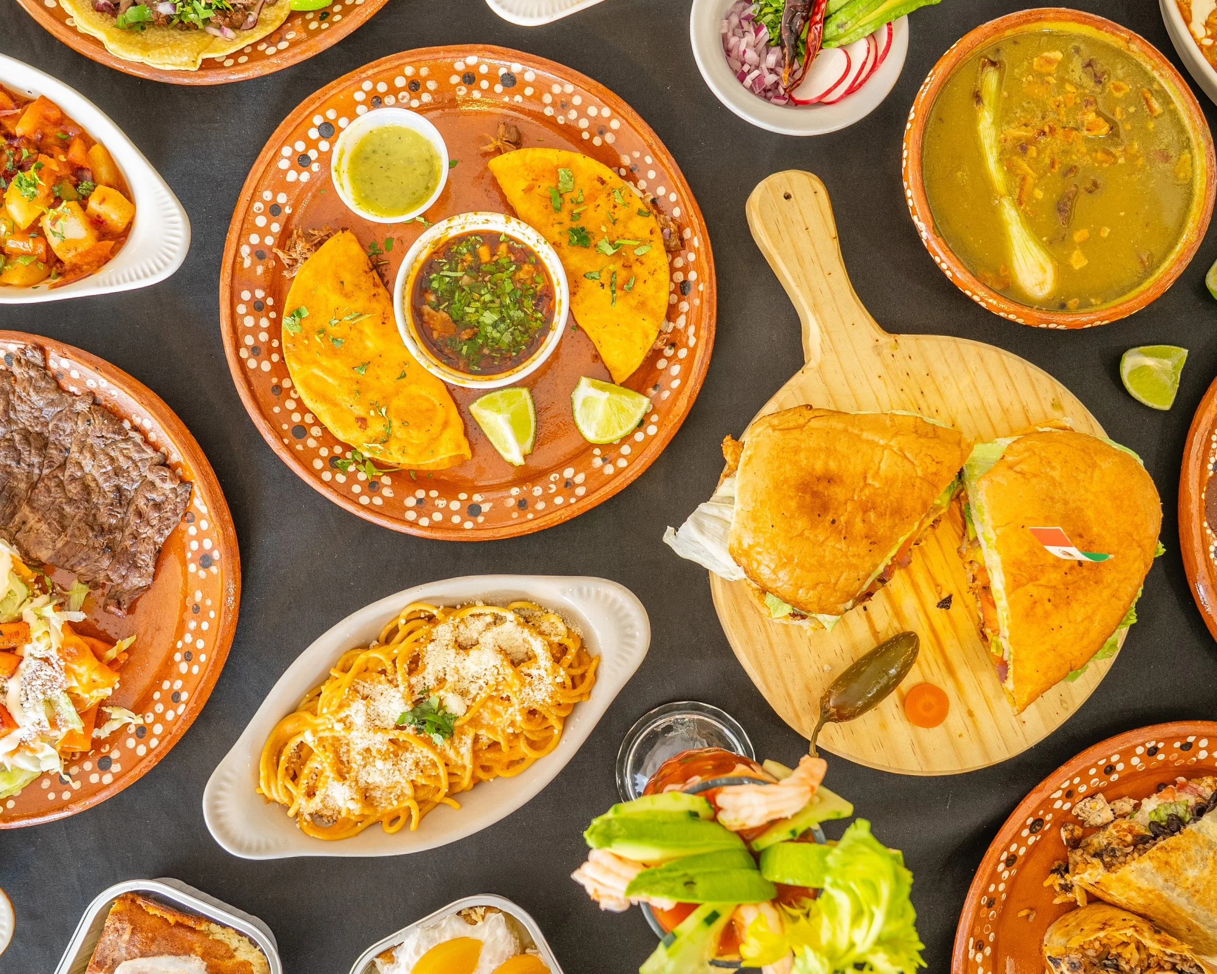 An overhead shot of various Mexican dishes including tacos, enchiladas, pasta, nachos, salsa, and salads served in different bowls and plates.