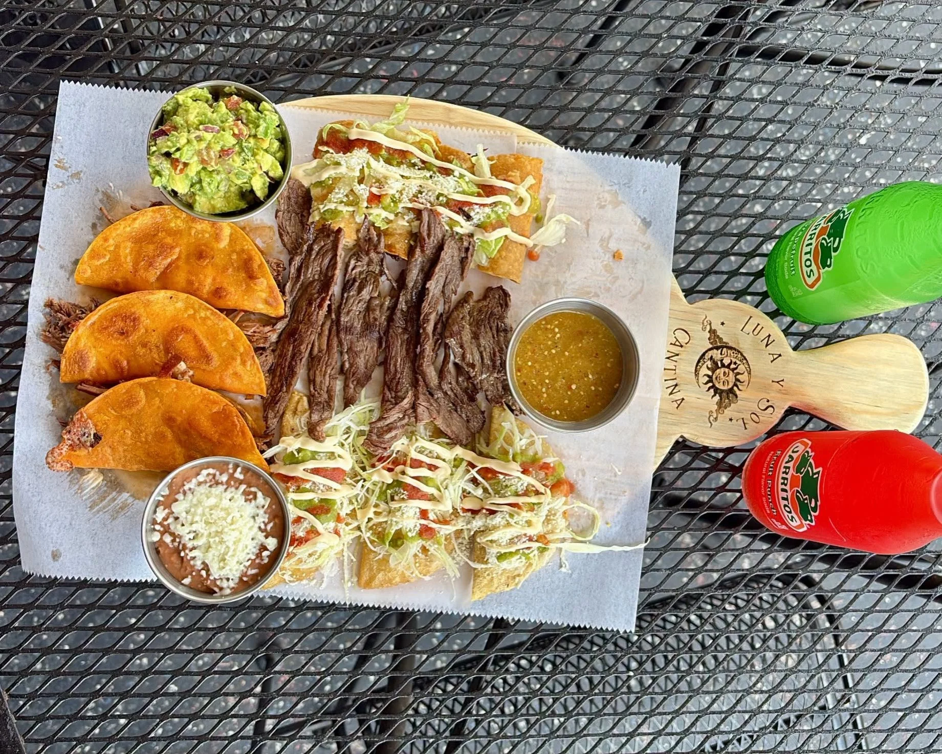 A Mexican meal on a metal outdoor table includes tacos, grilled beef steak, a side of guacamole, shredded cheese, and salsa in small containers. There are also two bottles of lime green and red flavored beverages.