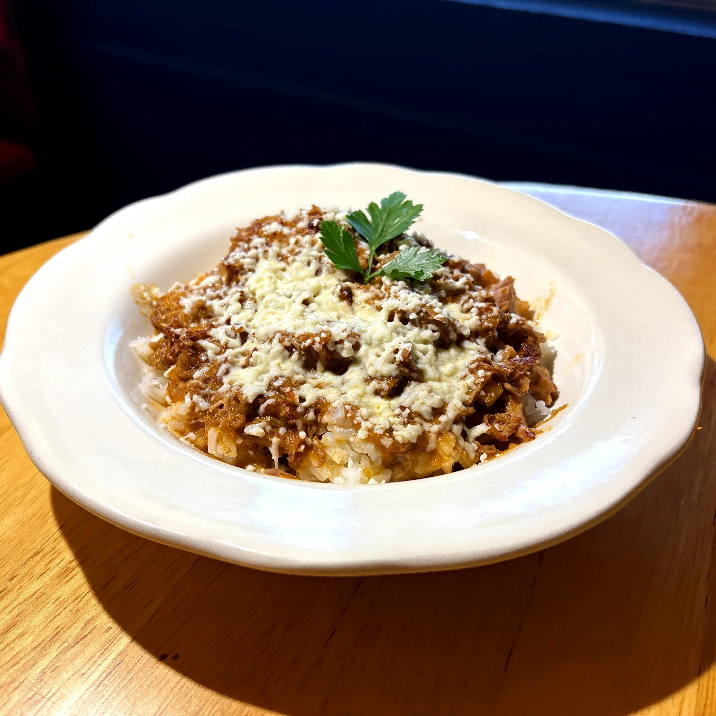 A white bowl of baked pasta topped with cheese and a sprig of parsley on a wooden table.