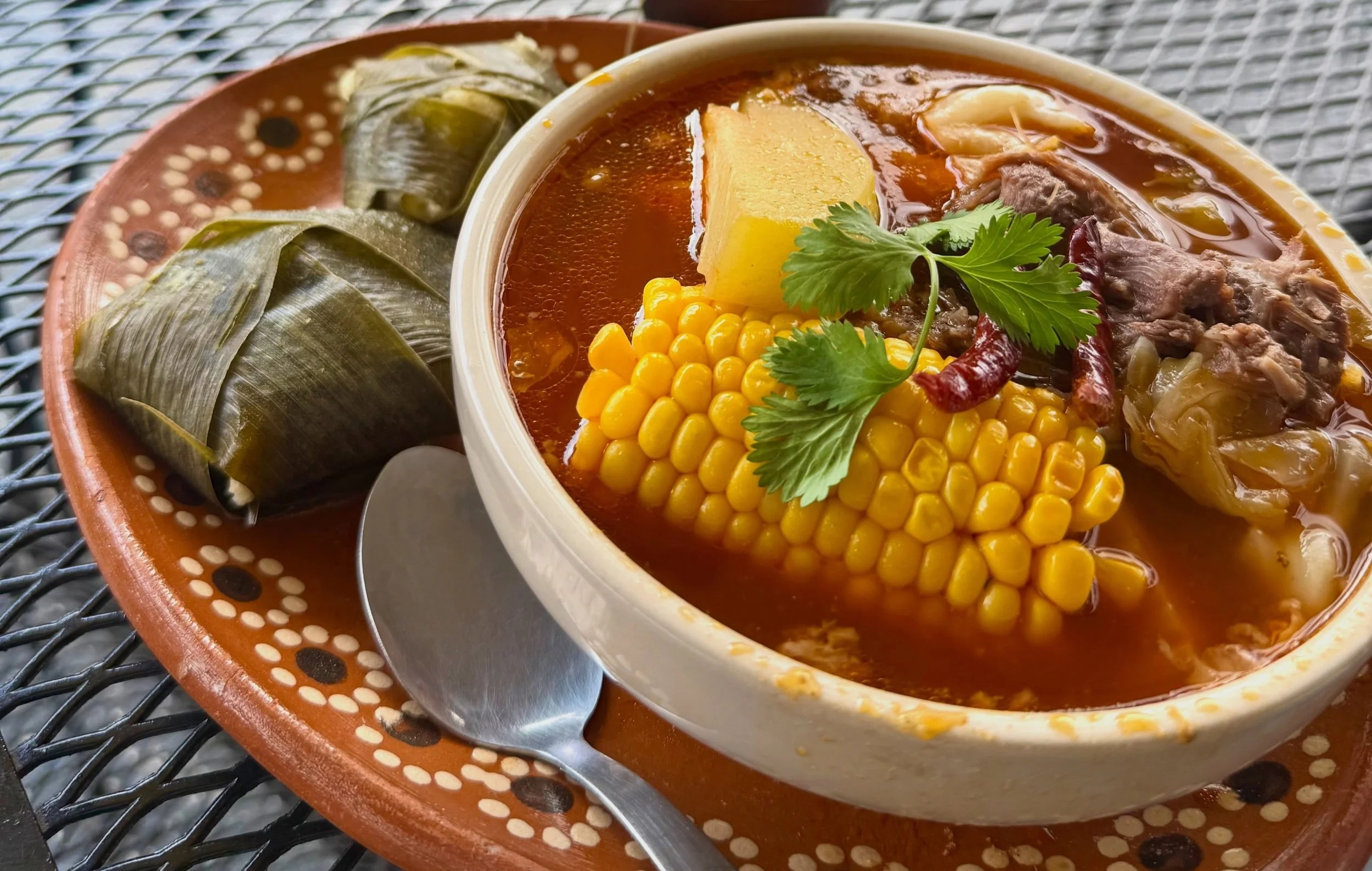 A bowl of hearty beef and vegetable soup garnished with fresh cilantro, accompanied by two tamales wrapped in corn husks, served on a decorative ceramic plate.