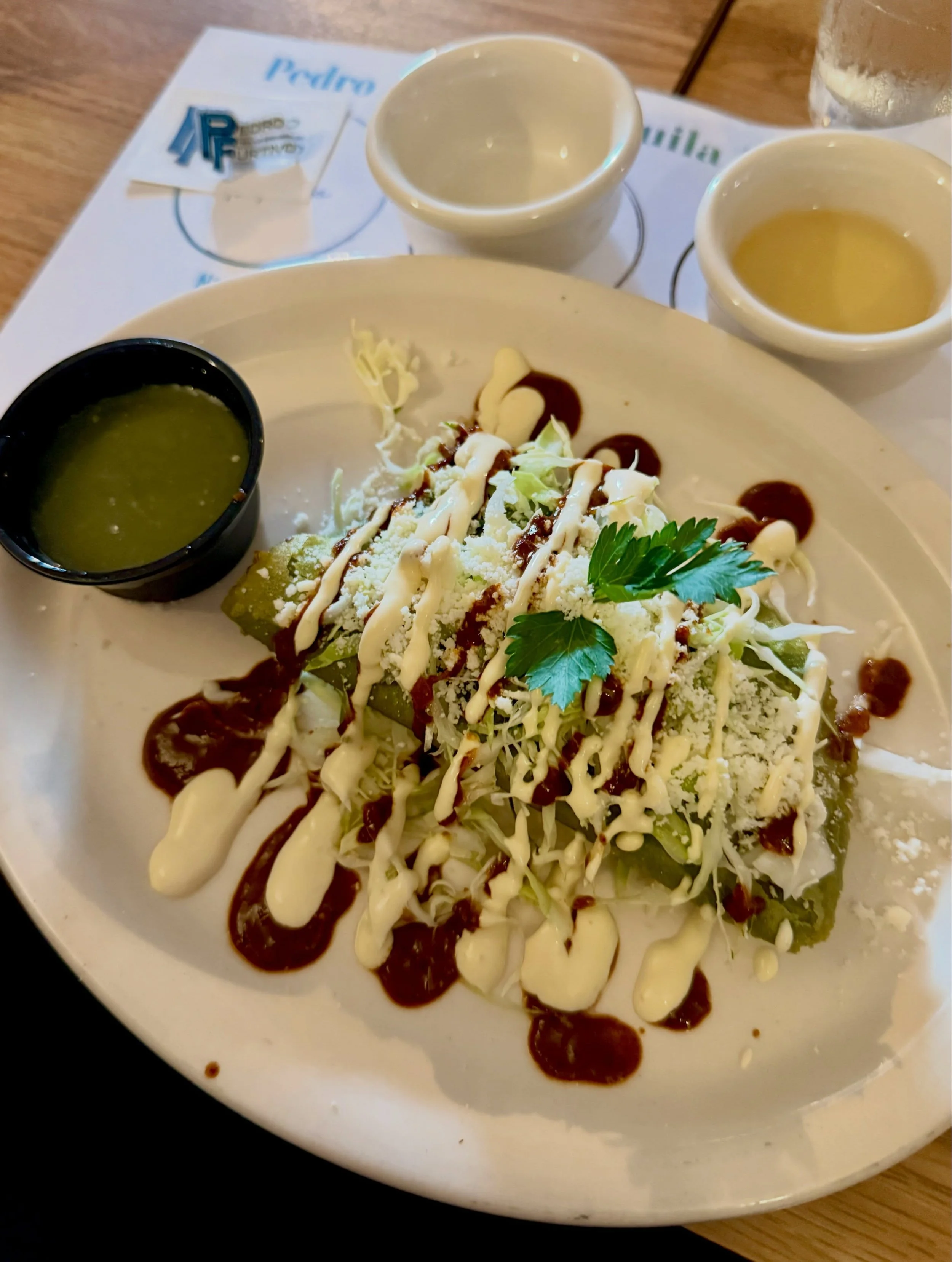 A plate of salad topped with shredded cheese, drizzled with creamy dressing and balsamic vinegar, garnished with a sprig of parsley, accompanied by small bowls of green dressing and another yellowish sauce or dressing, and a glass of water in the bac