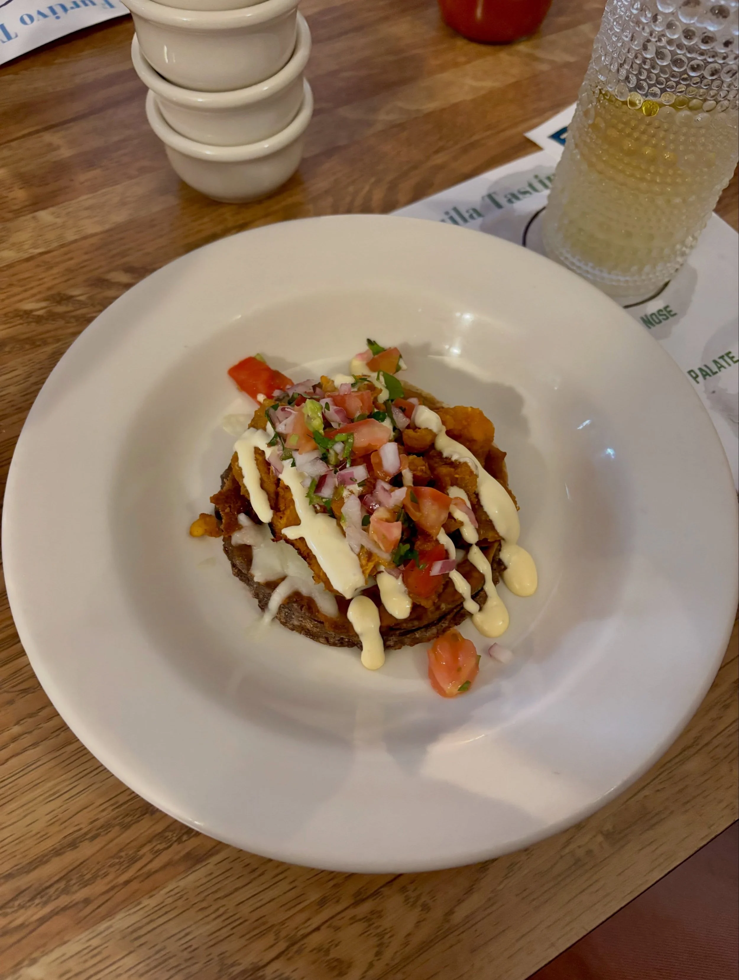 A plate of Mexican food featuring fried fish, topped with chopped tomatoes, jalapeños, and drizzled with creamy sauce, on a wooden table. Beside it, there is a glass of beer and small stacked bowls, with a napkin underneath the beer glass.