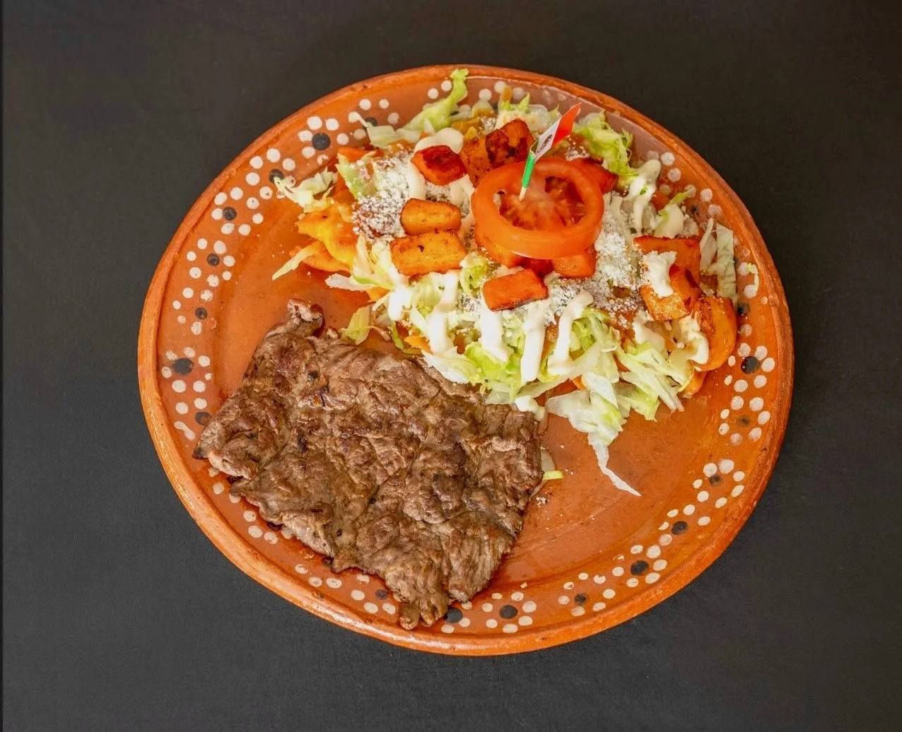 A plate of food with grilled beef, shredded lettuce, sliced tomato, croutons, and shredded cheese, served on a decorative orange plate against a black background.