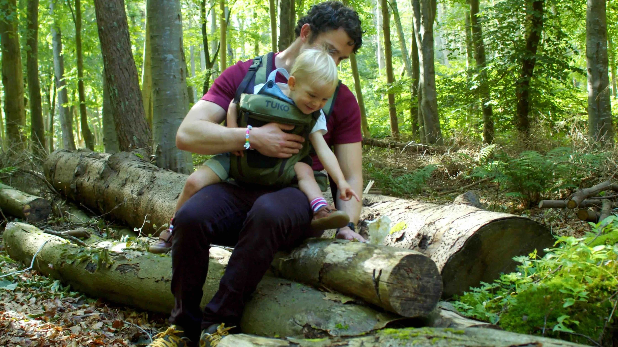 TUKO owner sitting on a forest log with his son secured in the TUKO baby carrier.